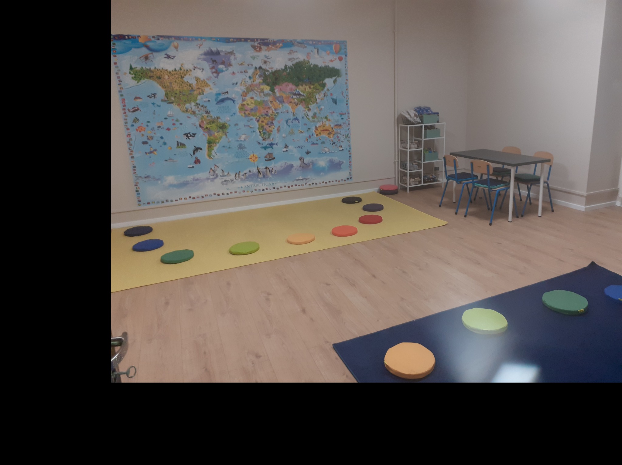 Jasne wnętrze sali zabaw dla dzieci z mapą świata na ścianie, kolorowymi poduszkami na podłodze i stolikiem z krzesłami w tle.