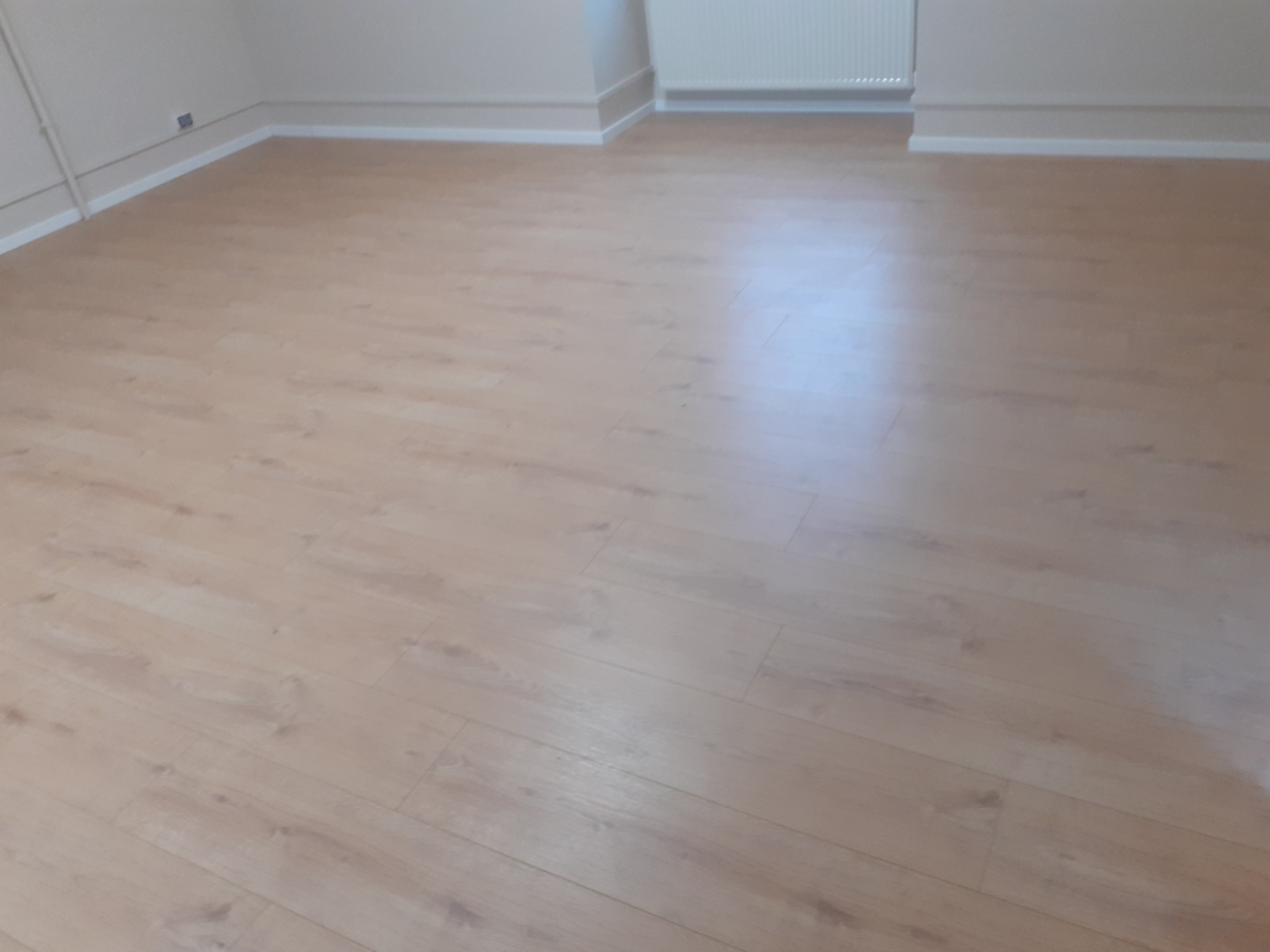 Jasne panele podłogowe ułożone w pustym pokoju z białymi listwami przypodłogowymi i grzejnikiem na ścianie.