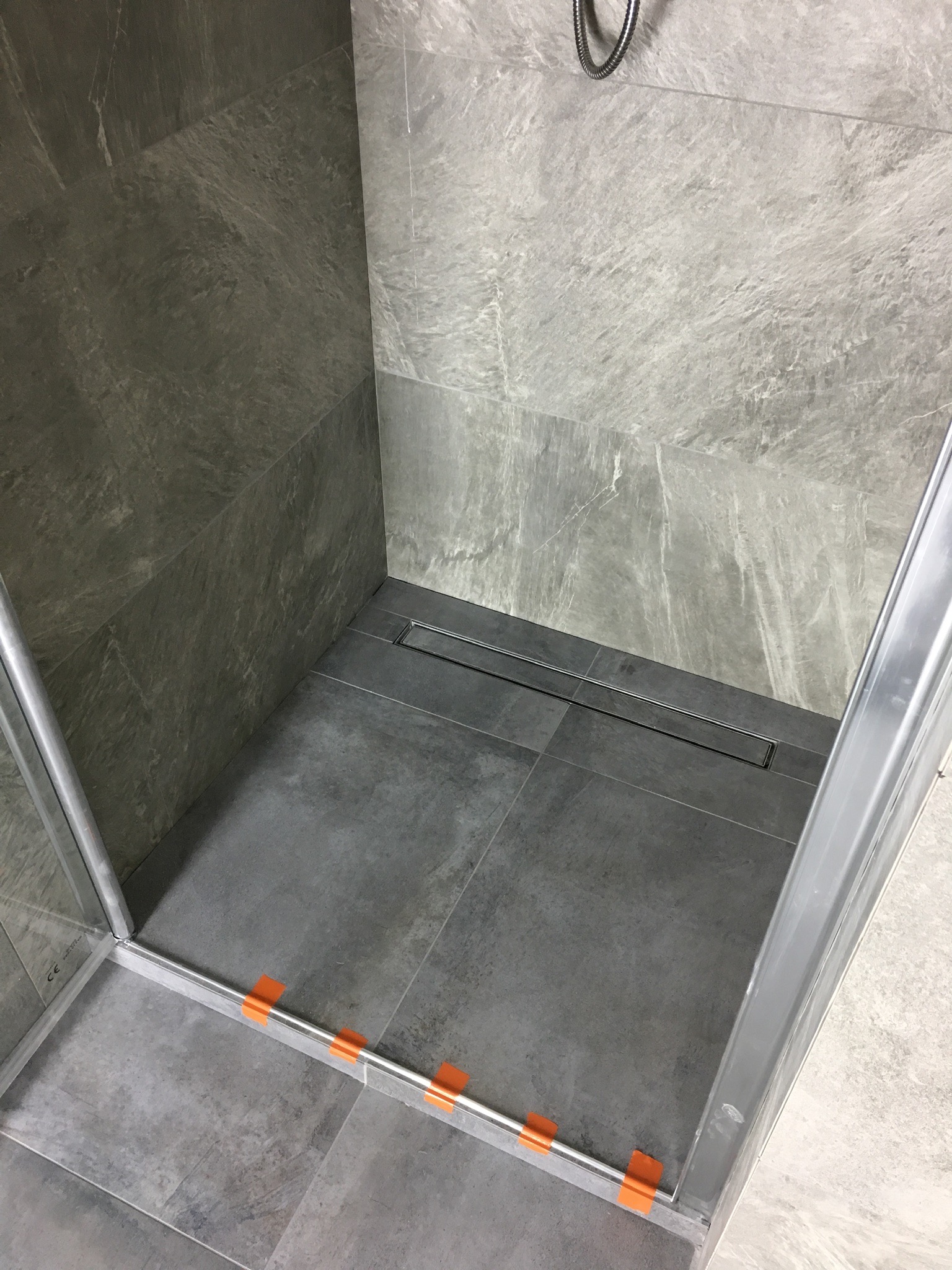 Nowoczesny prysznic walk-in z szarymi płytkami imitującymi kamień, liniowym odpływem i szklaną kabiną zabezpieczoną pomarańczową taśmą montażową.