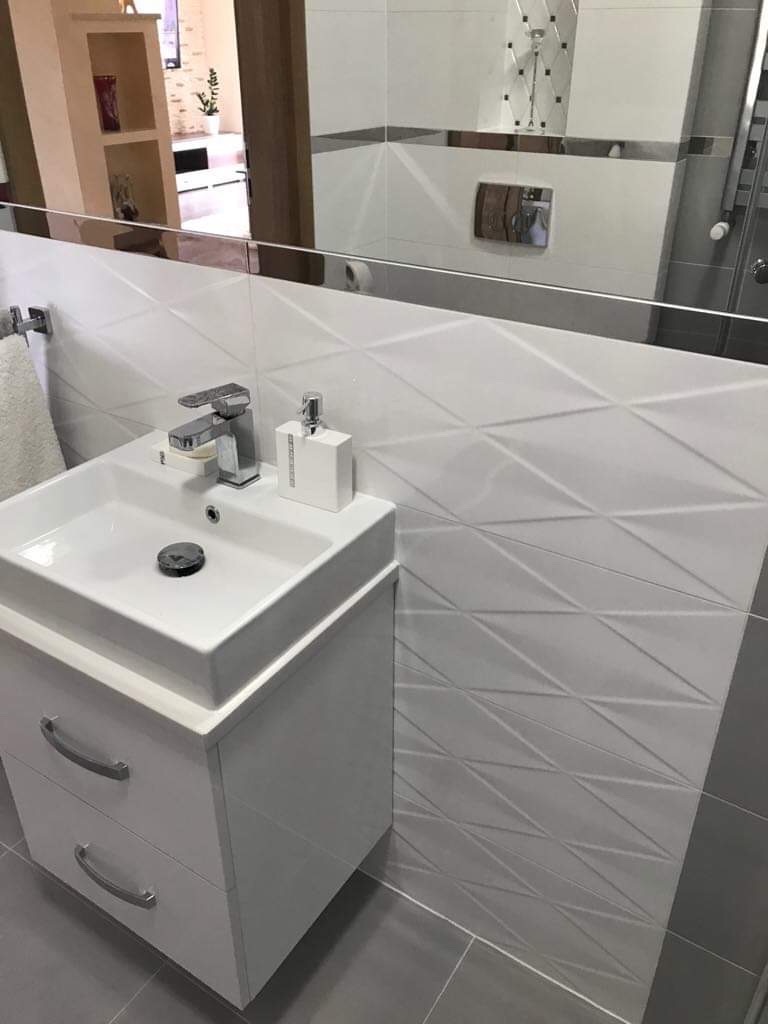 Nowoczesna łazienka z białą umywalką nablatową na szafce z szufladami, białe trójwymiarowe płytki na ścianie i szare płytki podłogowe, widoczne odbicie kabiny prysznicowej w lustrze.