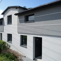 Dwukondygnacyjny dom po nałożeniu tynku zewnętrznego w odcieniach szarości, z widocznym fragmentem ogrodu i czarną rynną spustową.