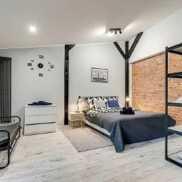 Jasne wnętrze sypialni z drewnianymi belkami pomalowanymi na czarno, zegarem ściennym z cyframi, łóżkiem z szarą narzutą i ceglaną ścianą w tle.