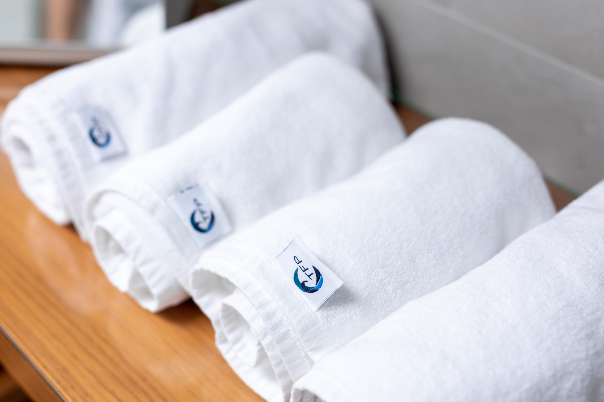 Rząd czterech zwiniętych białych ręczników frotte z logo TFP na drewnianym blacie, widoczne odbicie w tle.