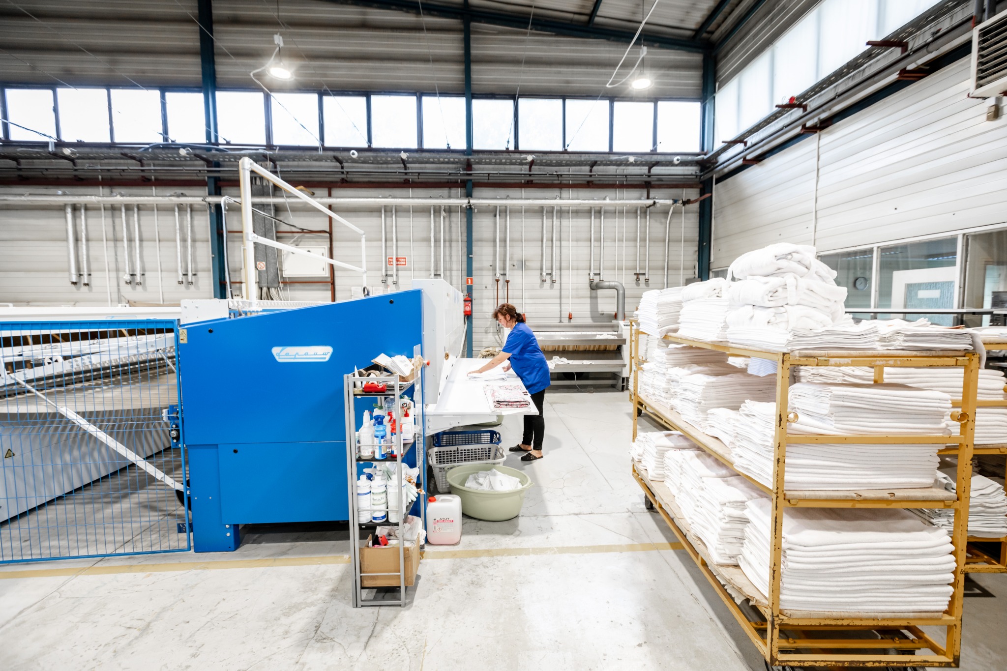 Pracownica w niebieskiej bluzie obsługuje magiel przemysłowy w pralni, obok wózki z ułożonymi ręcznikami i pościelą, widoczny regał z detergentami i środkami czystości.