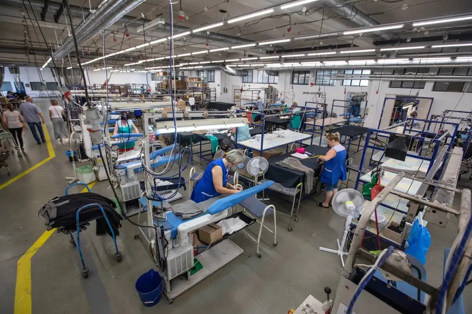 Duża hala produkcyjna z pracownikami przy stanowiskach prasowania i kontroli jakości odzieży, widoczne liczne stoły, maszyny do prasowania z wytwornicami pary, wentylatory, w tle osoby poruszające...