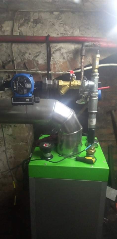 Nowoczesny piec z zielonym blatem i srebrną obudową, podłączony do instalacji z widoczną pompą obiegową, rurami miedzianymi i zaworami, na tle ściany z surowej cegły.