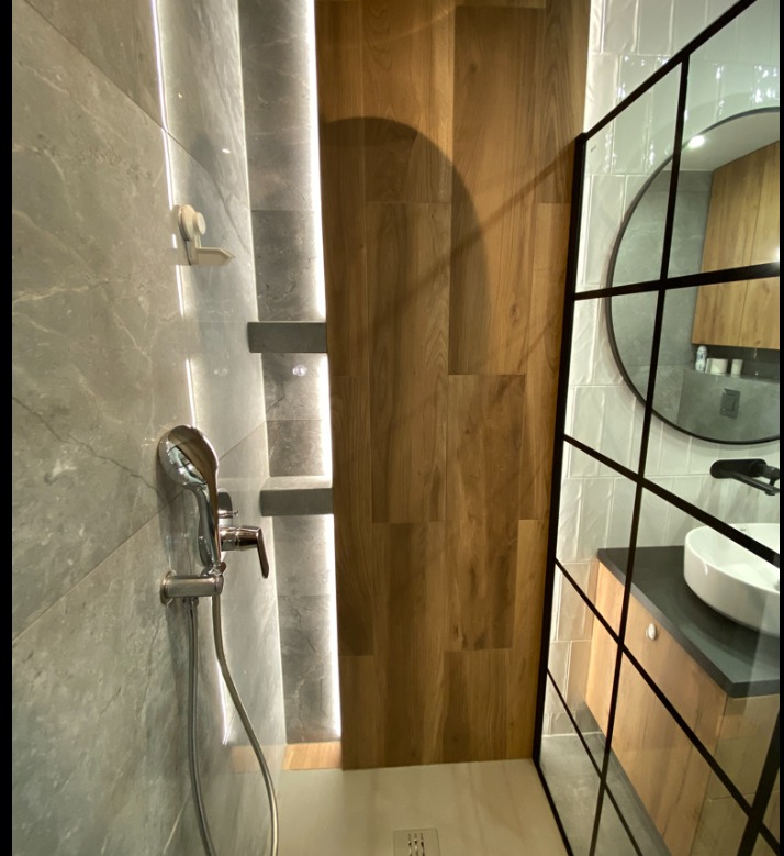Nowoczesna łazienka w Gdańsku z kabiną prysznicową typu walk-in, wykończona szarymi płytkami imitującymi kamień i panelami drewnopodobnymi, z oświetleniem LED wzdłuż ściany prysznicowej i okrągłym...