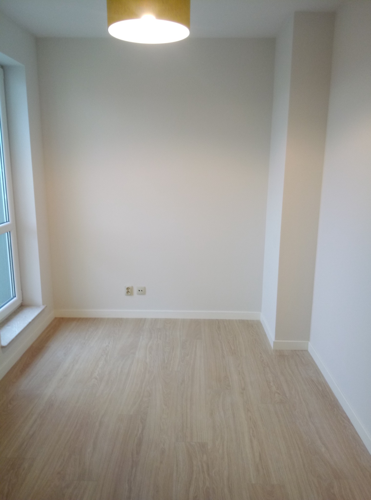 Pusty pokój z panelami podłogowymi w odcieniach beżu, świeżo pomalowanymi białymi ścianami, widoczne gniazdka elektryczne i żółta lampa sufitowa.