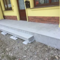 Wyłożone jasnoszarą płytką schody wejściowe do domu z żółtą elewacją i brązowymi elementami dekoracyjnymi, widoczne podpory pod schodami na żwirowym podłożu.