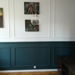 Ściana z panelami pomalowanymi na kolor ciemnego turkusu, białe panele g&oacute;rne, parkiet ułożony w jodełkę, wisząca lampa z przezroczystego szkła, obrazy na ścianie.