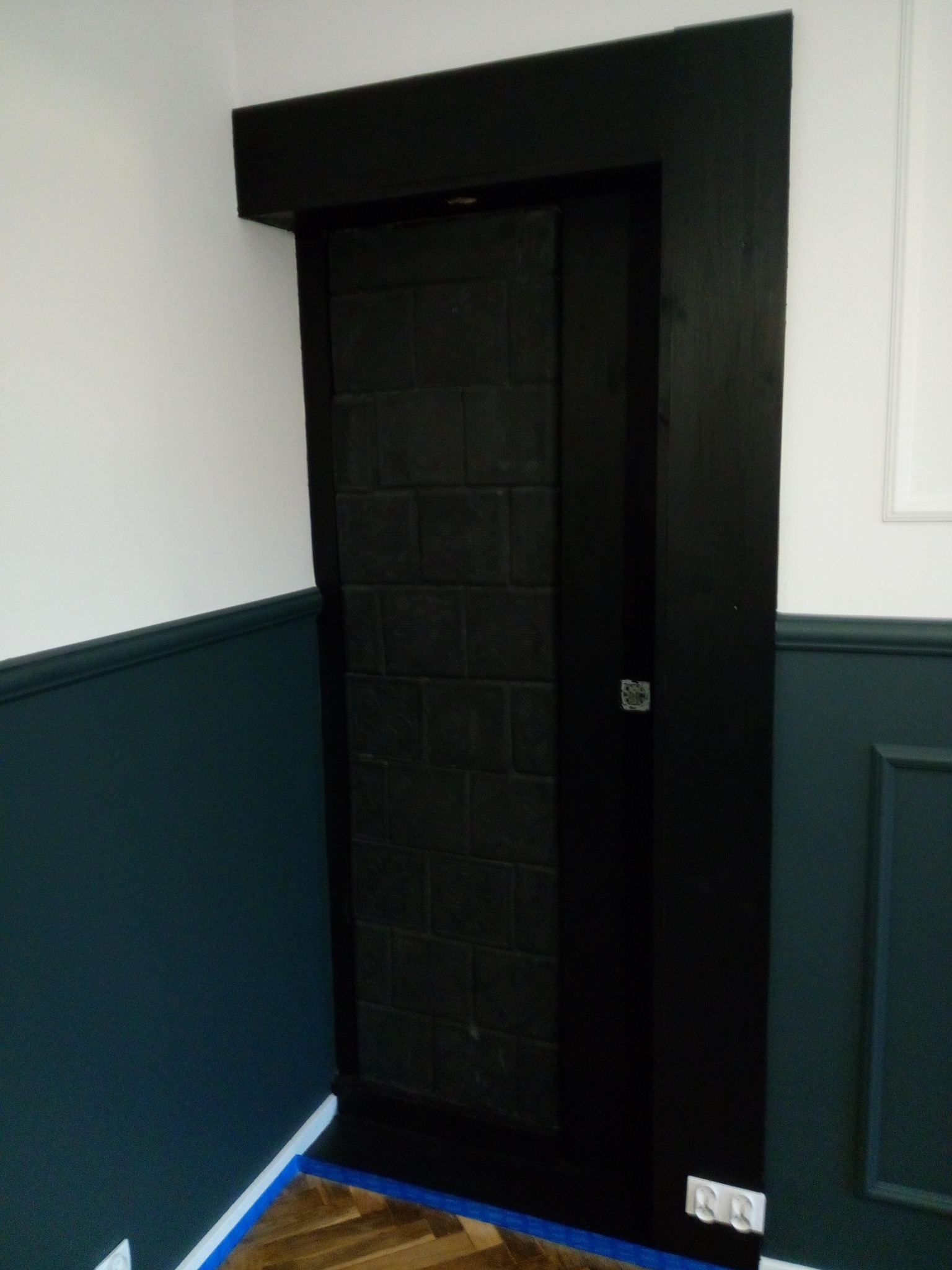 Narożne ujęcie czarnych, ceglanych drzwi ukrytych w ścianie, otoczonych czarną ramą, z widocznymi gniazdkami elektrycznymi u dołu i niebieską taśmą malarską na podłodze.