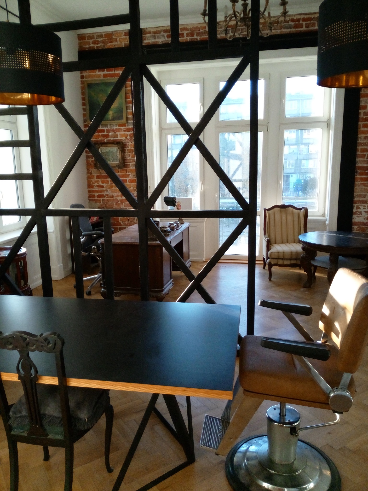 Loftowe biuro z czarnym biurkiem, krzesłem fryzjerskim i ścianą z czerwonej cegły, widok przez metalową konstrukcję.