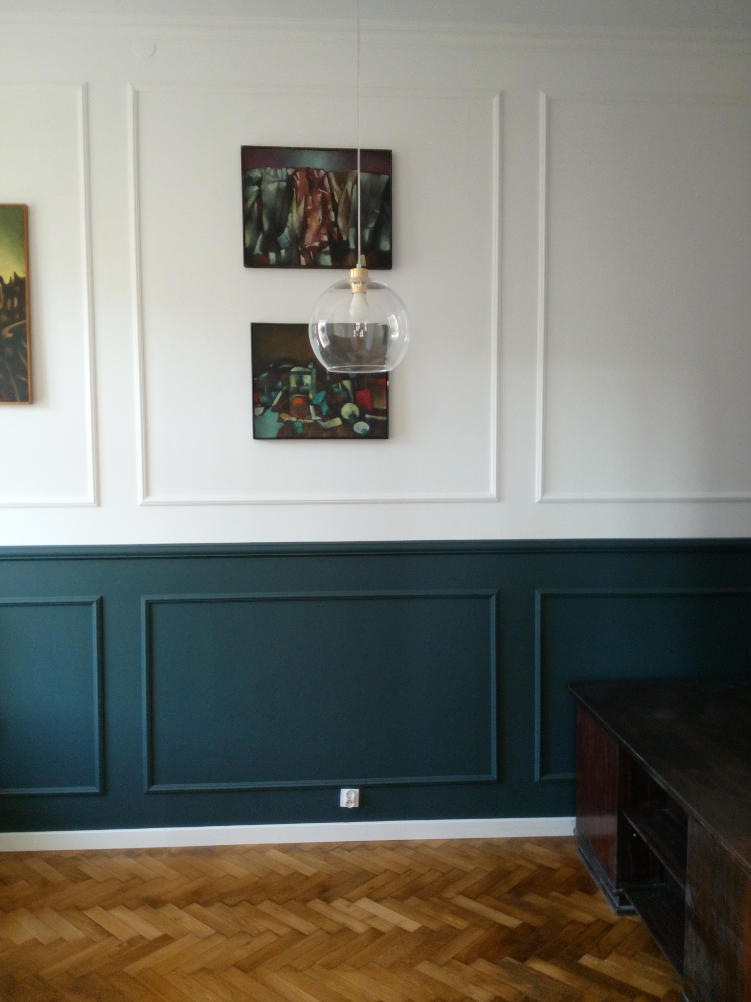 Ściana z panelami pomalowanymi na kolor ciemnego turkusu, białe panele górne, parkiet ułożony w jodełkę, wisząca lampa z przezroczystego szkła, obrazy na ścianie.