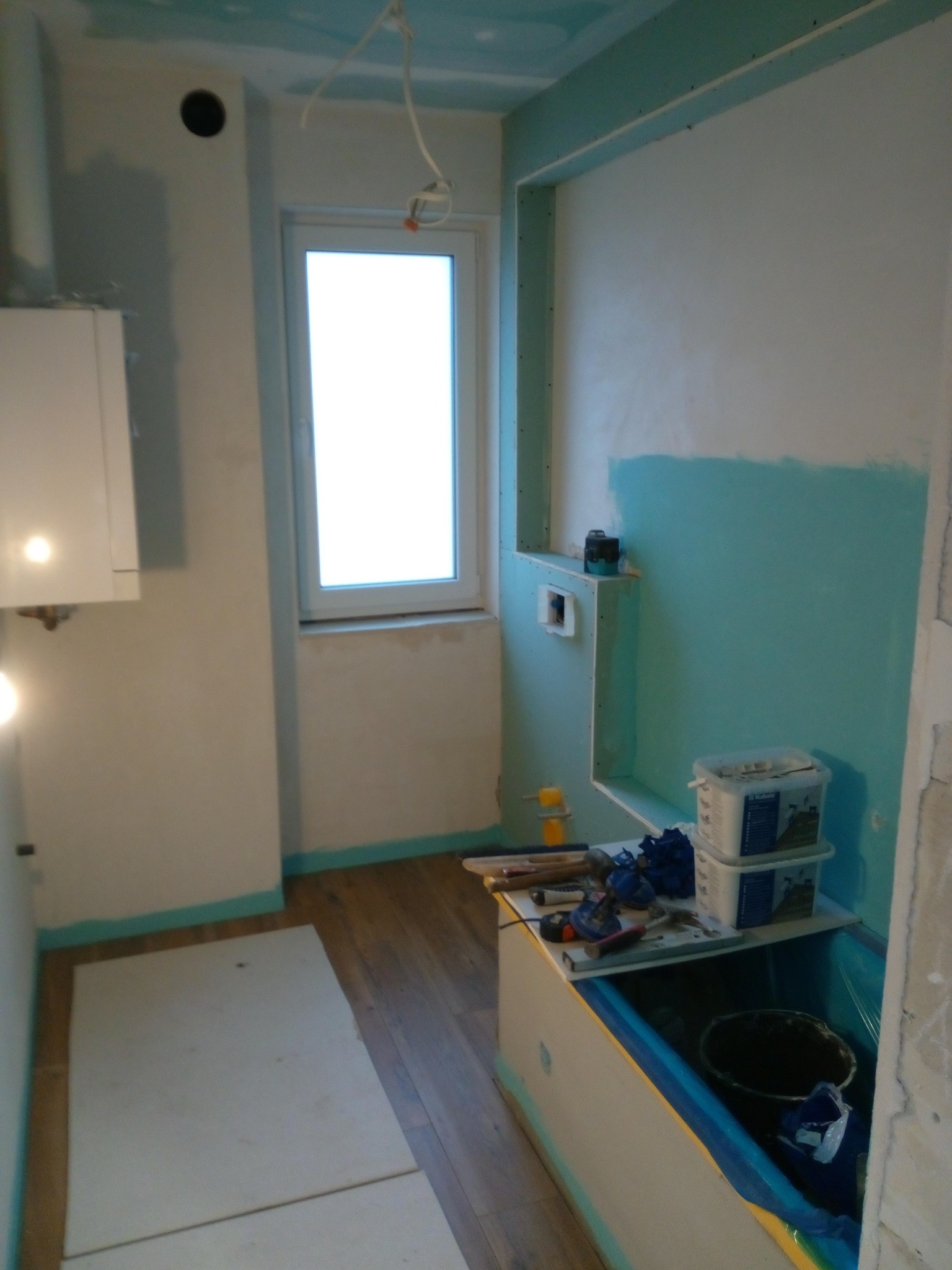 Łazienka w trakcie remontu: częściowo pomalowane ściany na turkusowo, widoczne narzędzia i materiały budowlane, zabudowa wanny, płyta karton gips na suficie.