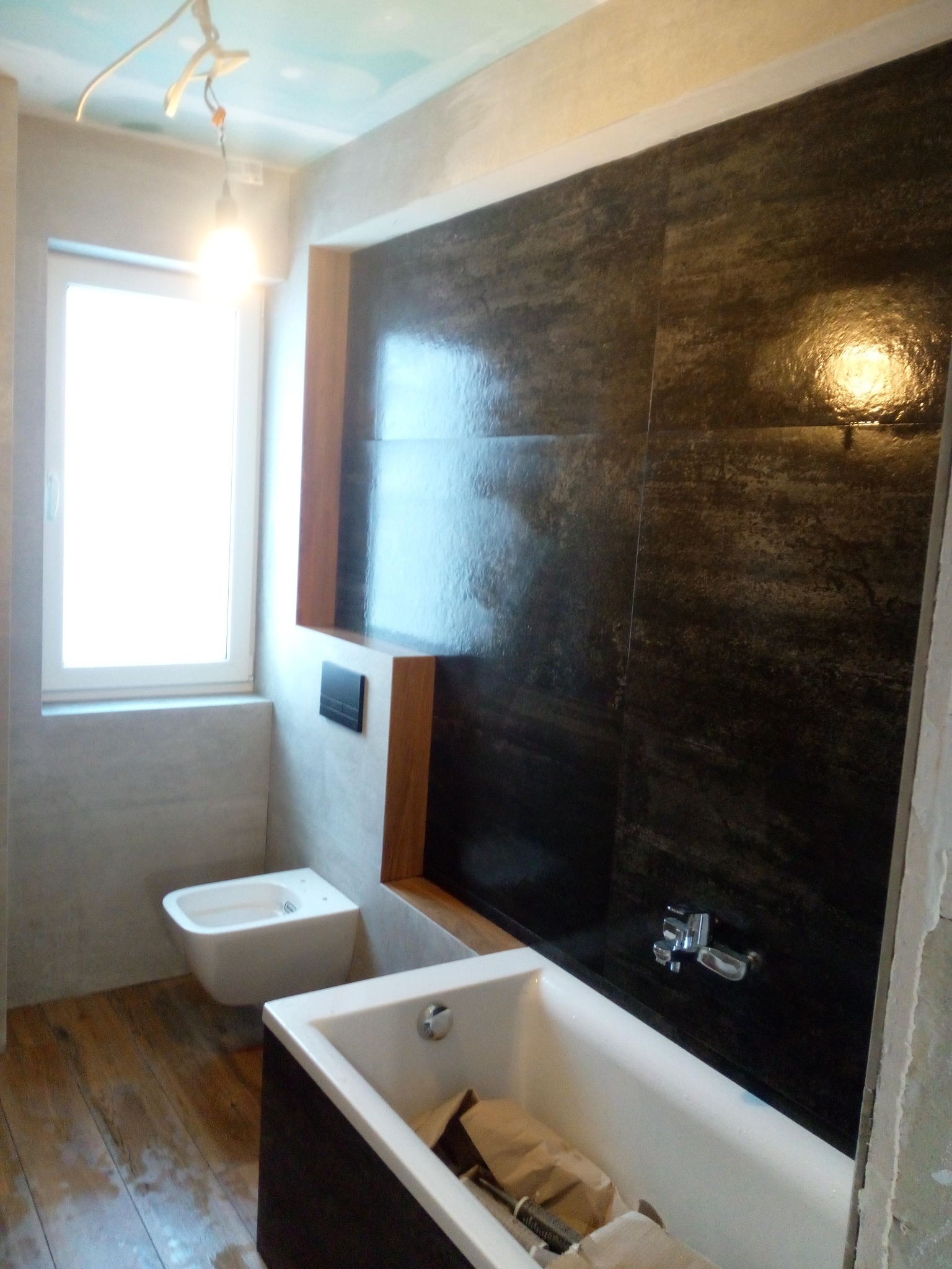 Niedokończona łazienka z ciemnymi, błyszczącymi płytkami na ścianie, białą wanną z brązowym obramowaniem, bidetem i drewnianą podłogą; surowy sufit z wiszącą żarówką.