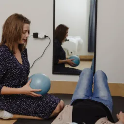 Kobieta w granatowej sukience z białymi kropkami siedzi na podłodze i trzyma niebieską piłkę, pacjentka leży na macie na plecach, widoczne nogi w jeansach, w tle lustro i regał z akcesoriami.