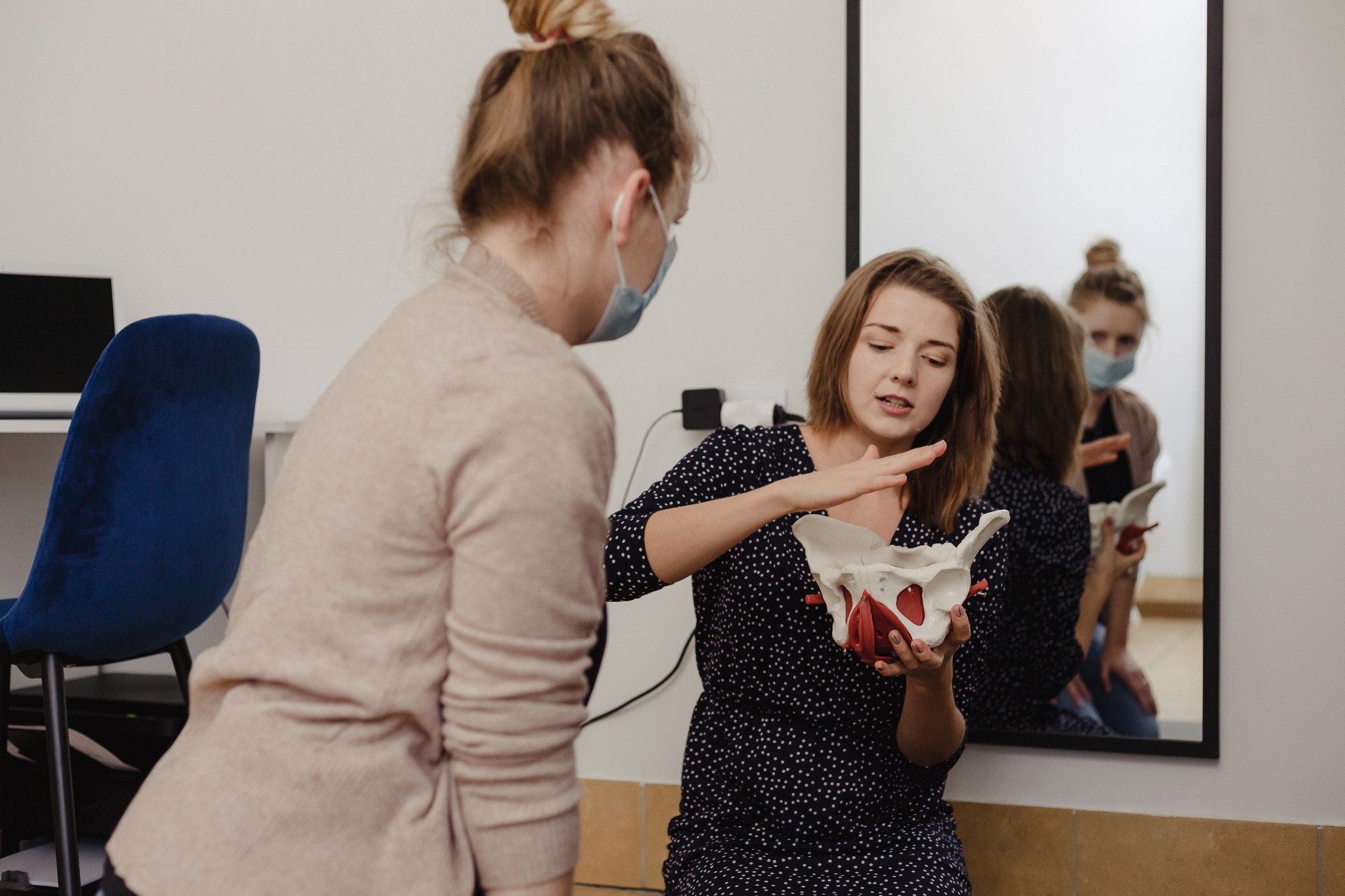 Kobieta w granatowej sukience w białe kropki demonstruje model miednicy pacjentce w beżowym swetrze i maseczce, odbicie w lustrze ukazuje obie kobiety.