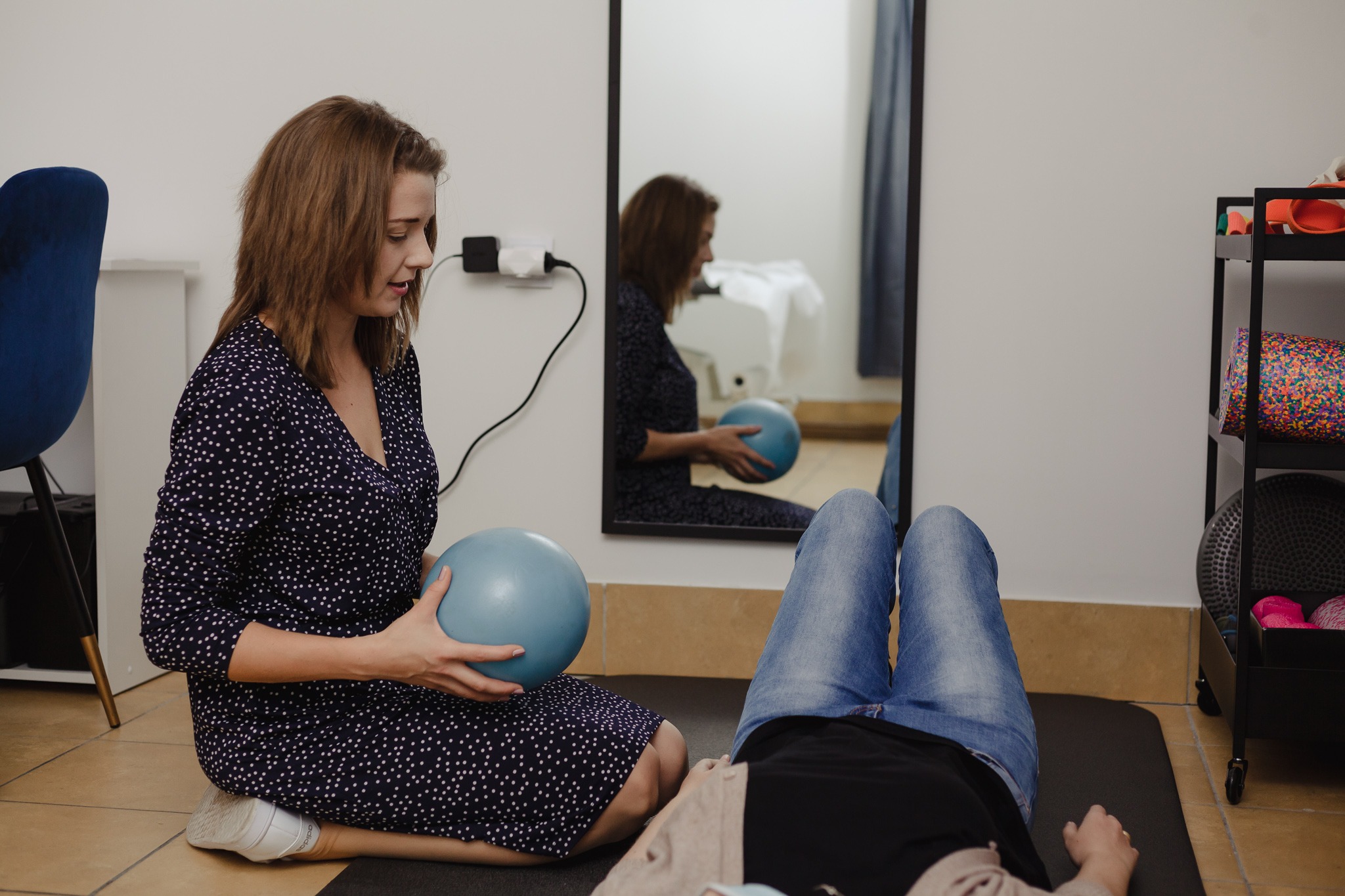 Kobieta w granatowej sukience z białymi kropkami siedzi na podłodze i trzyma niebieską piłkę, pacjentka leży na macie na plecach, widoczne nogi w jeansach, w tle lustro i regał z akcesoriami.