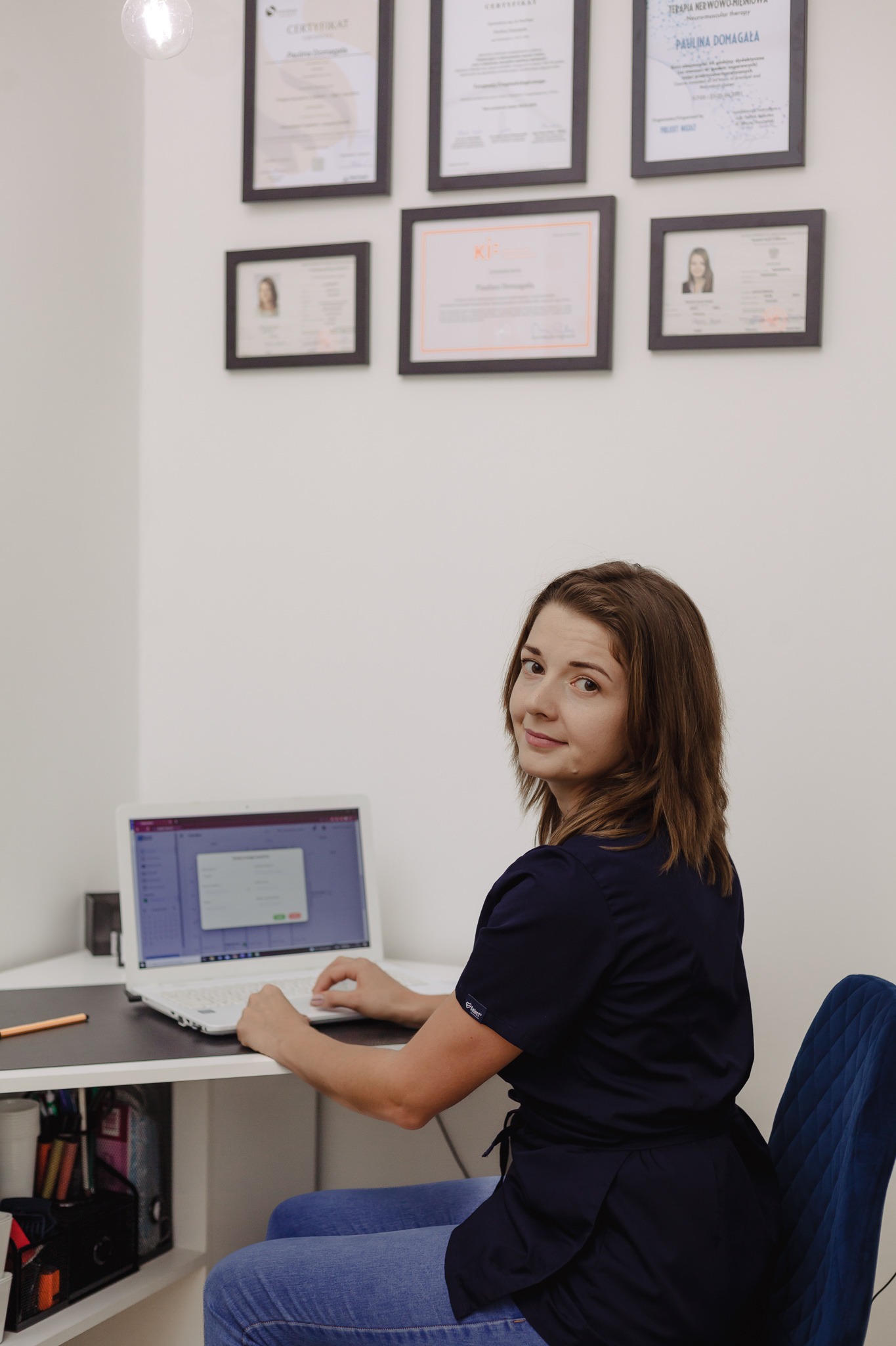 Kobieta w granatowym uniformie siedzi przy biurku z laptopem, odwrócona w stronę kamery, na ścianie wiszą certyfikaty i dyplomy w ramkach.