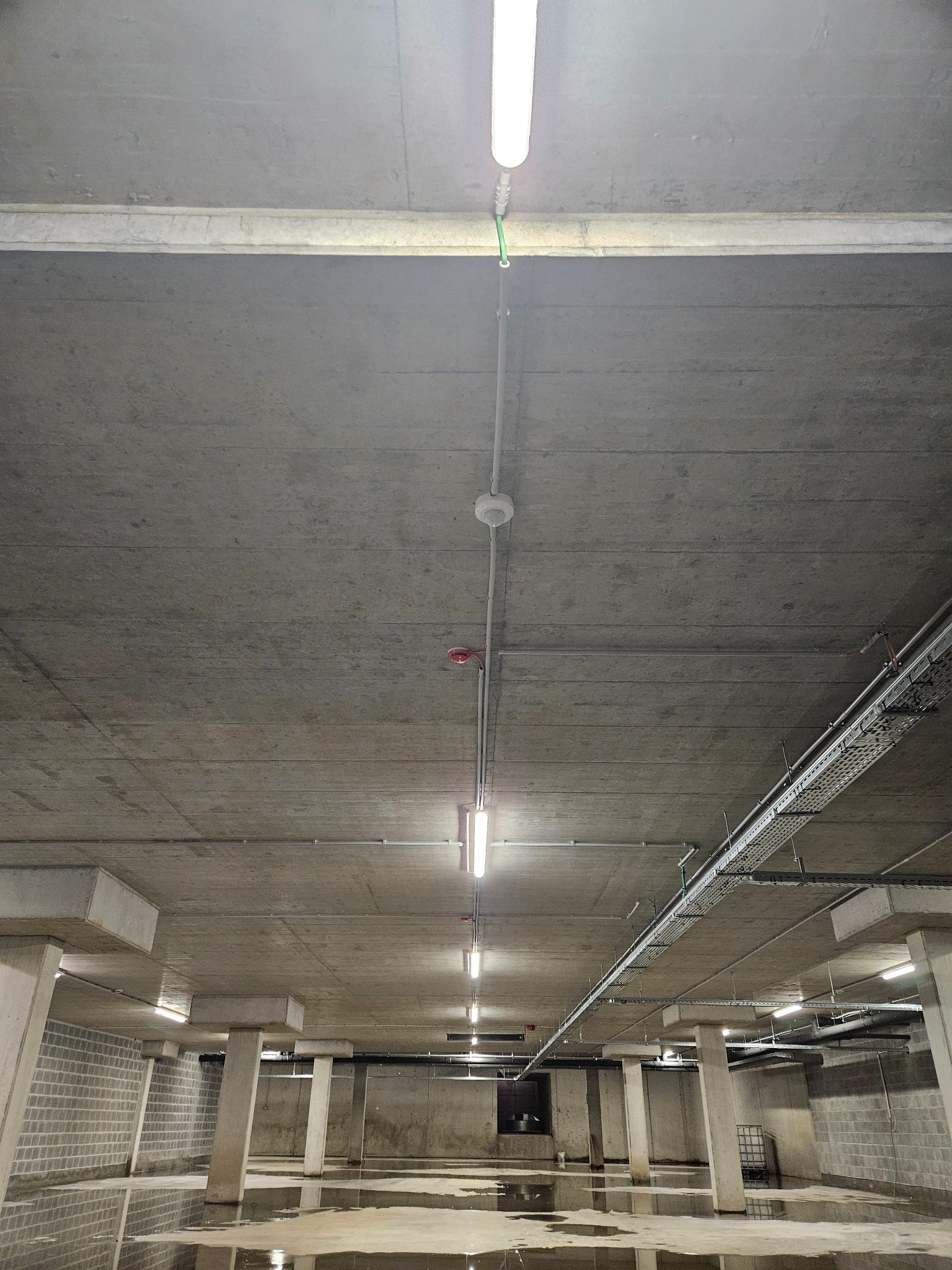 Wnętrze betonowego garażu podziemnego z widocznymi instalacjami elektrycznymi pod sufitem, w tym lampami, przewodami i czujnikiem dymu. Na podłodze stojąca woda. Widoczne kolumny podtrzymujące strop.