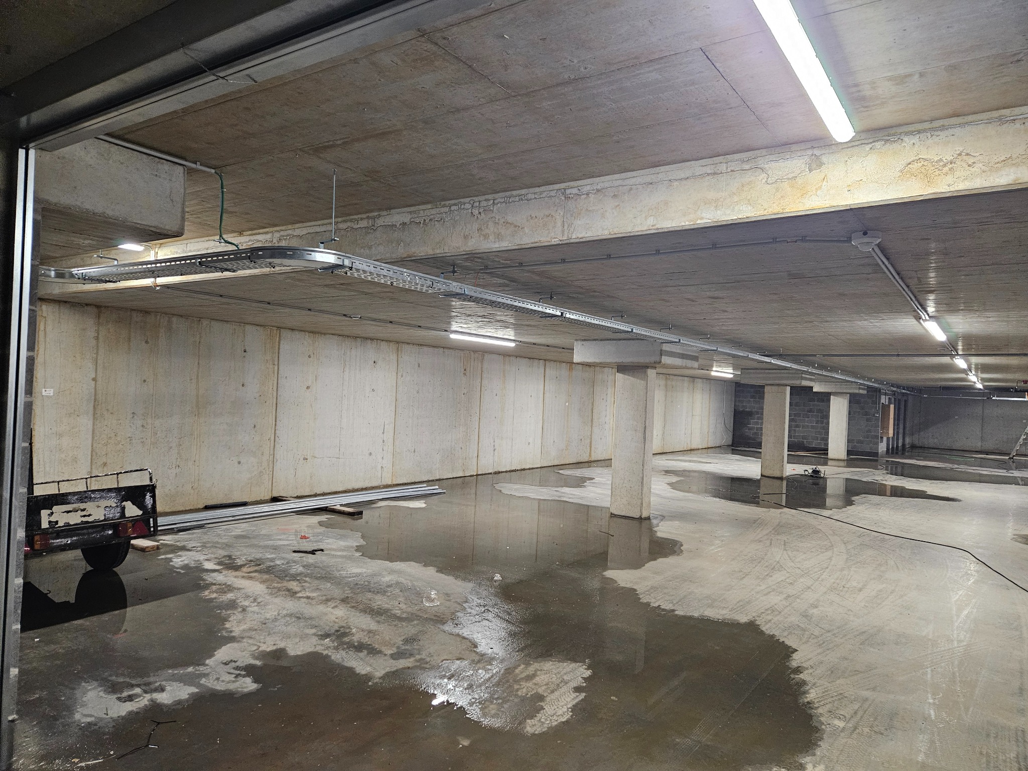 Instalacja koryta kablowego pod sufitem w surowym wnętrzu parkingu podziemnego z betonowymi ścianami i mokrą posadzką, widoczne rury instalacyjne i oświetlenie LED.