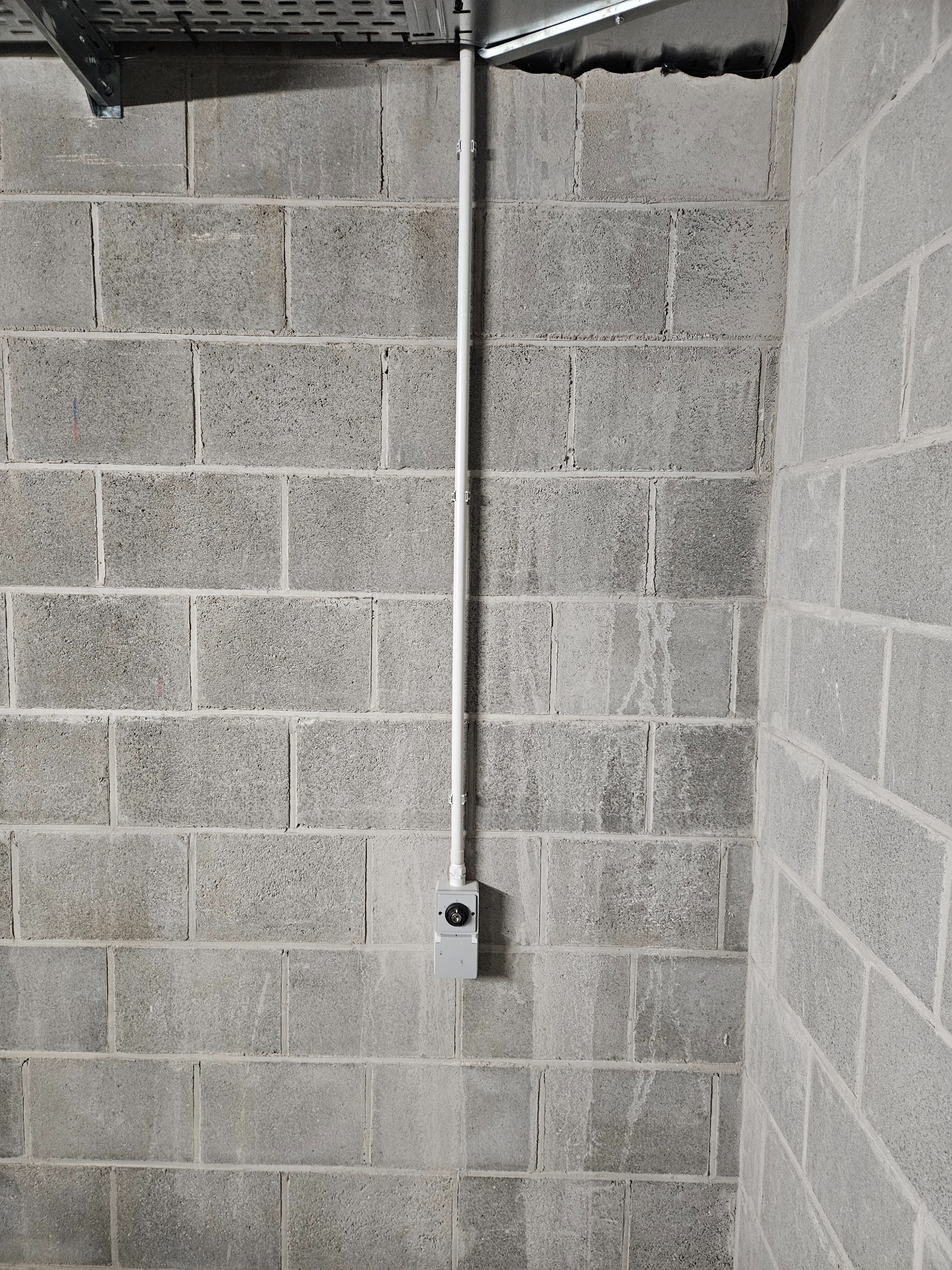 Szara ściana z bloków betonowych, na której pionowo biegnie biała rura instalacyjna z szarą puszką i czarnym włącznikiem na wysokości wzroku, widoczny fragment metalowej konstrukcji pod sufitem.
