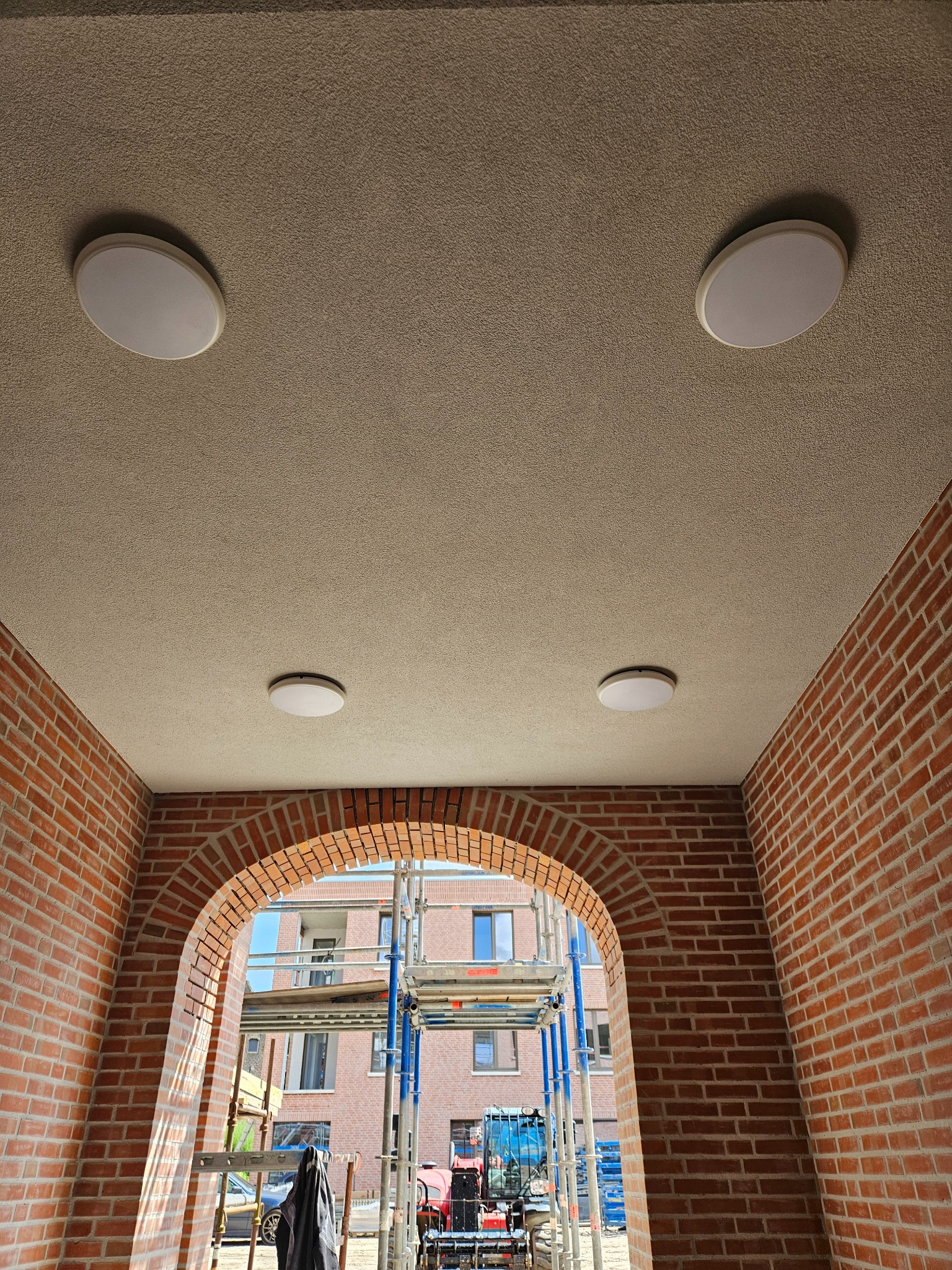 Cztery okrągłe lampy sufitowe zamontowane na teksturowanym suficie w przejściu z ceglanym łukiem, na zewnątrz widoczne rusztowania i budynek.