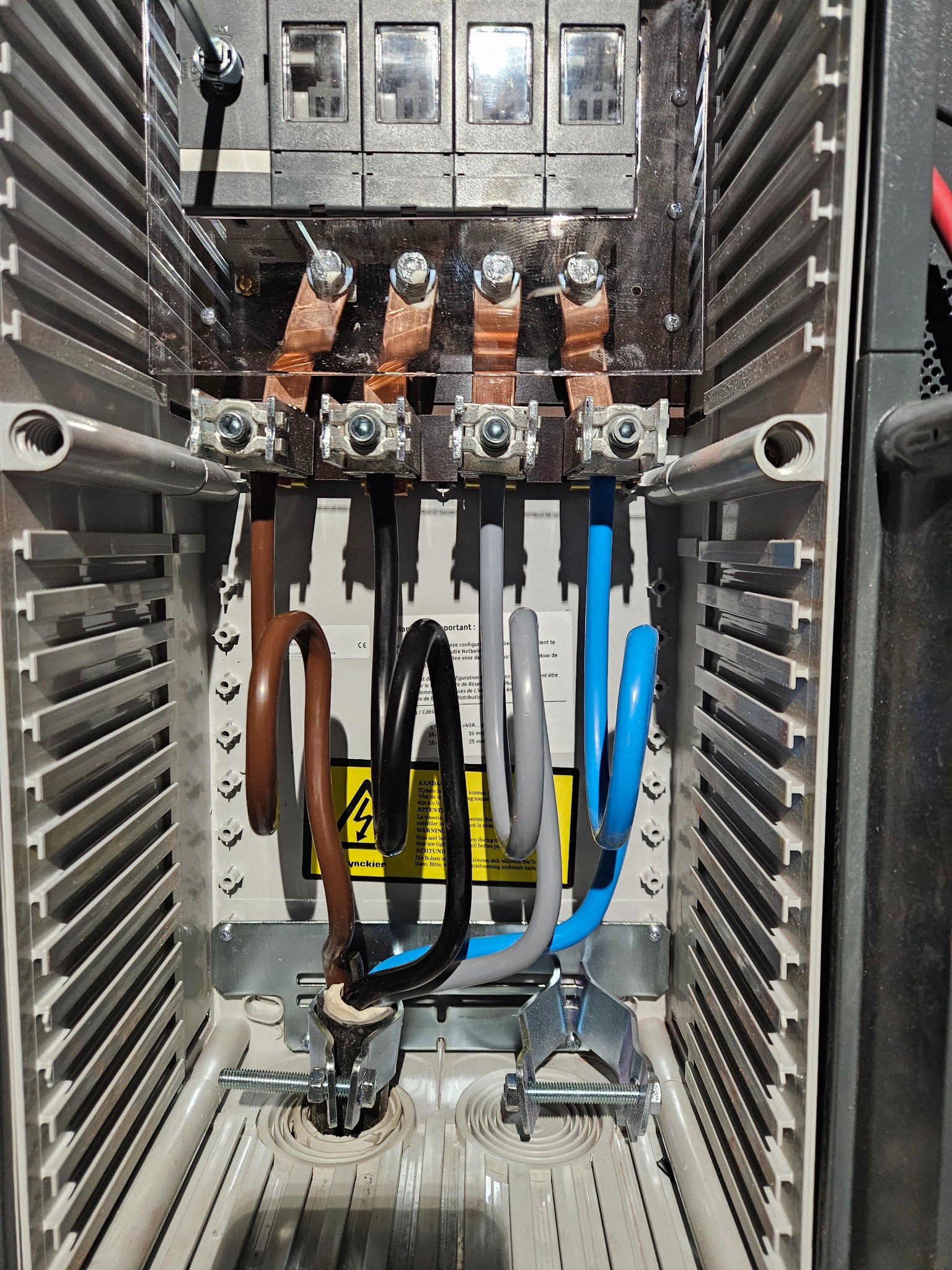 Rozdzielnica elektryczna z widocznymi przewodami w kolorach brązowym, czarnym, szarym i niebieskim, podłączonymi do górnych zacisków miedzianych; przewody ułożone w pętle, zamocowane do dolnych...