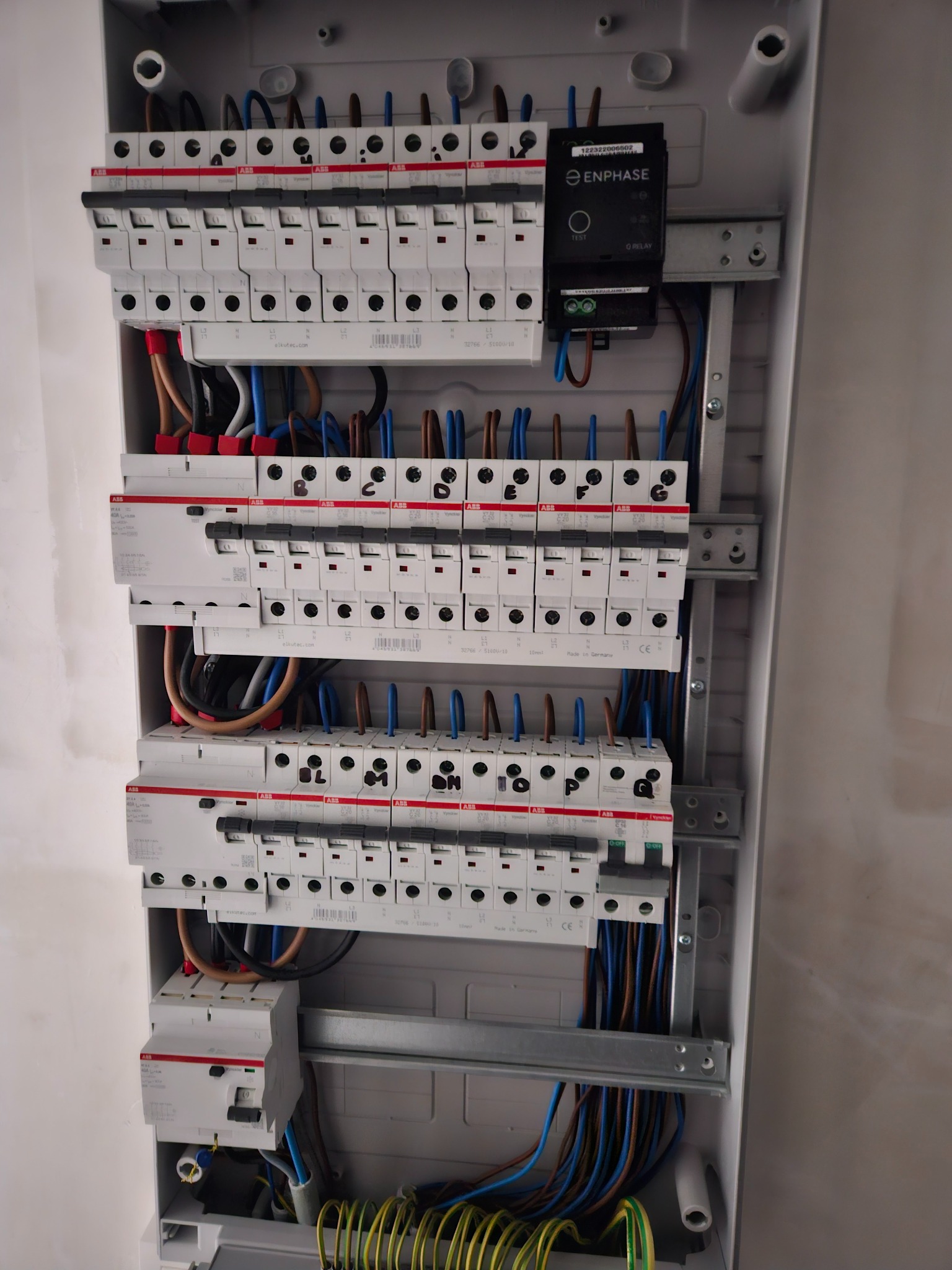 Rozdzielnica elektryczna z zainstalowanymi bezpiecznikami automatycznymi ABB i modułem Enphase, widoczne przewody w różnych kolorach, szyna montażowa i elementy mocujące wewnątrz obudowy z tworzywa