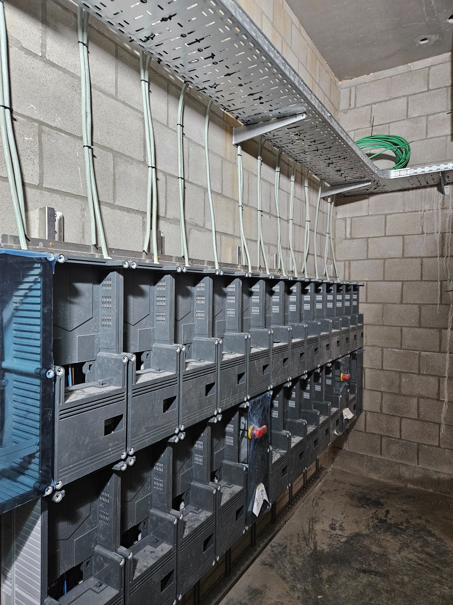Szara, betonowa ściana z zamontowanymi korytkami kablowymi i rozdzielnicami elektrycznymi; widoczne przewody w peszlach.