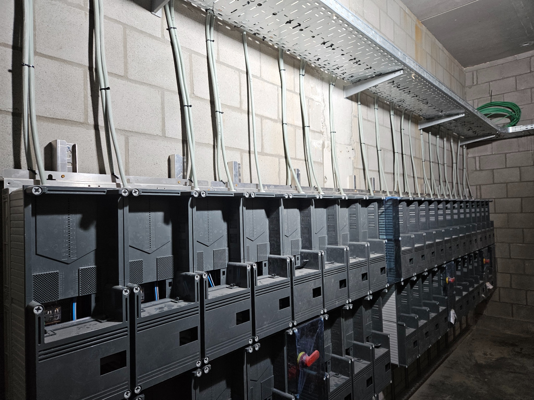 Szereg szarych skrzynek elektrycznych zamontowanych na ścianie z cegły, z przewodami biegnącymi w metalowej korycie kablowej pod sufitem.