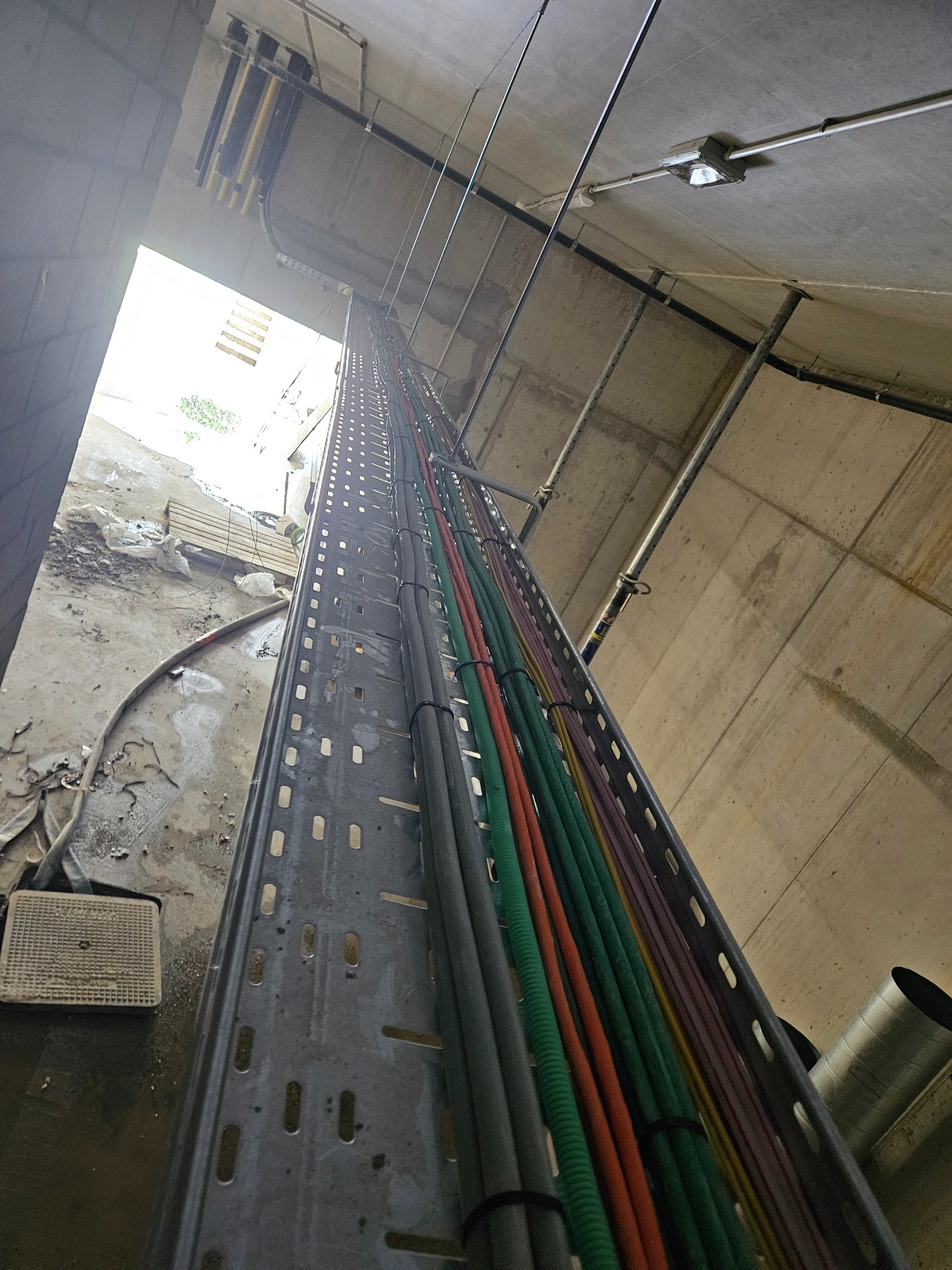 Metalowa korytka kablowa z kolorowymi przewodami elektrycznymi, zamocowana pod betonowym sufitem w surowym wnętrzu budynku w trakcie instalacji.