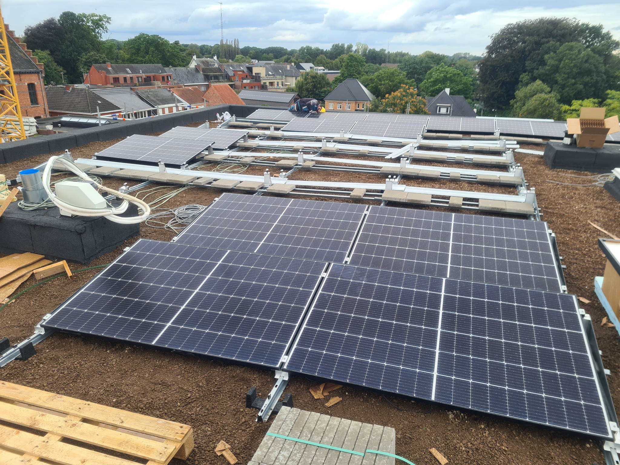 Montaż paneli słonecznych na płaskim dachu budynku mieszkalnego, widoczny pracownik w kasku, elementy montażowe, kable i skrzynka przyłączeniowa, w tle zabudowa miejska i drzewa.