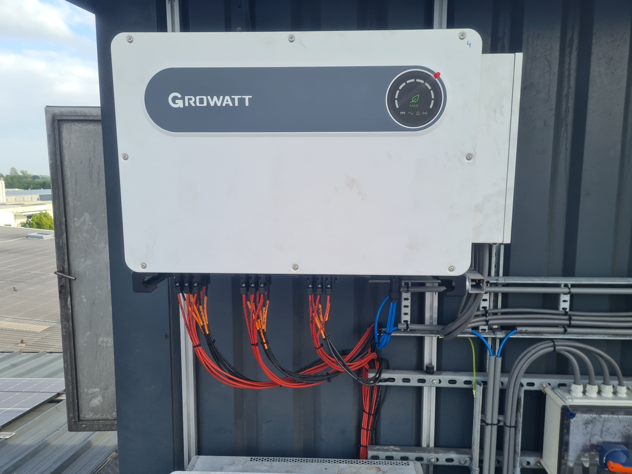 Biały falownik Growatt zamontowany na ciemnej ścianie, widoczne kable w kolorach czarnym, czerwonym i pomarańczowym, elementy instalacji na dachu w tle.