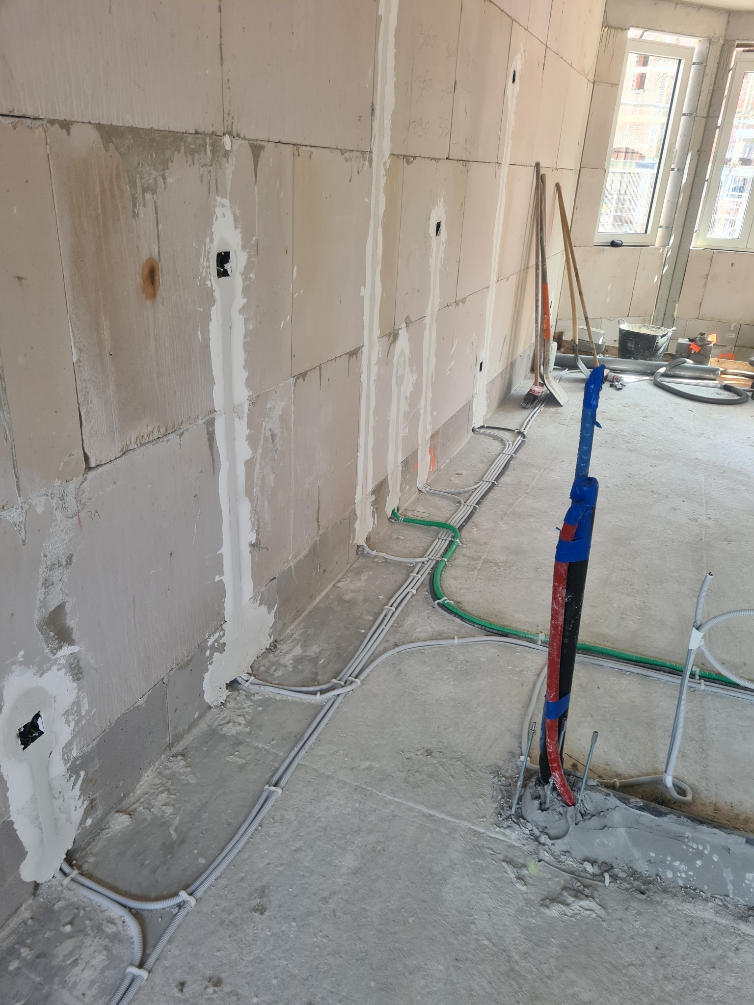 Surowy stan wnętrza z widocznymi przewodami elektrycznymi w listwach przypodłogowych, pionowymi rurami zabezpieczonymi niebieską taśmą, częściowo otynkowanymi ścianami z widocznymi puszkami...