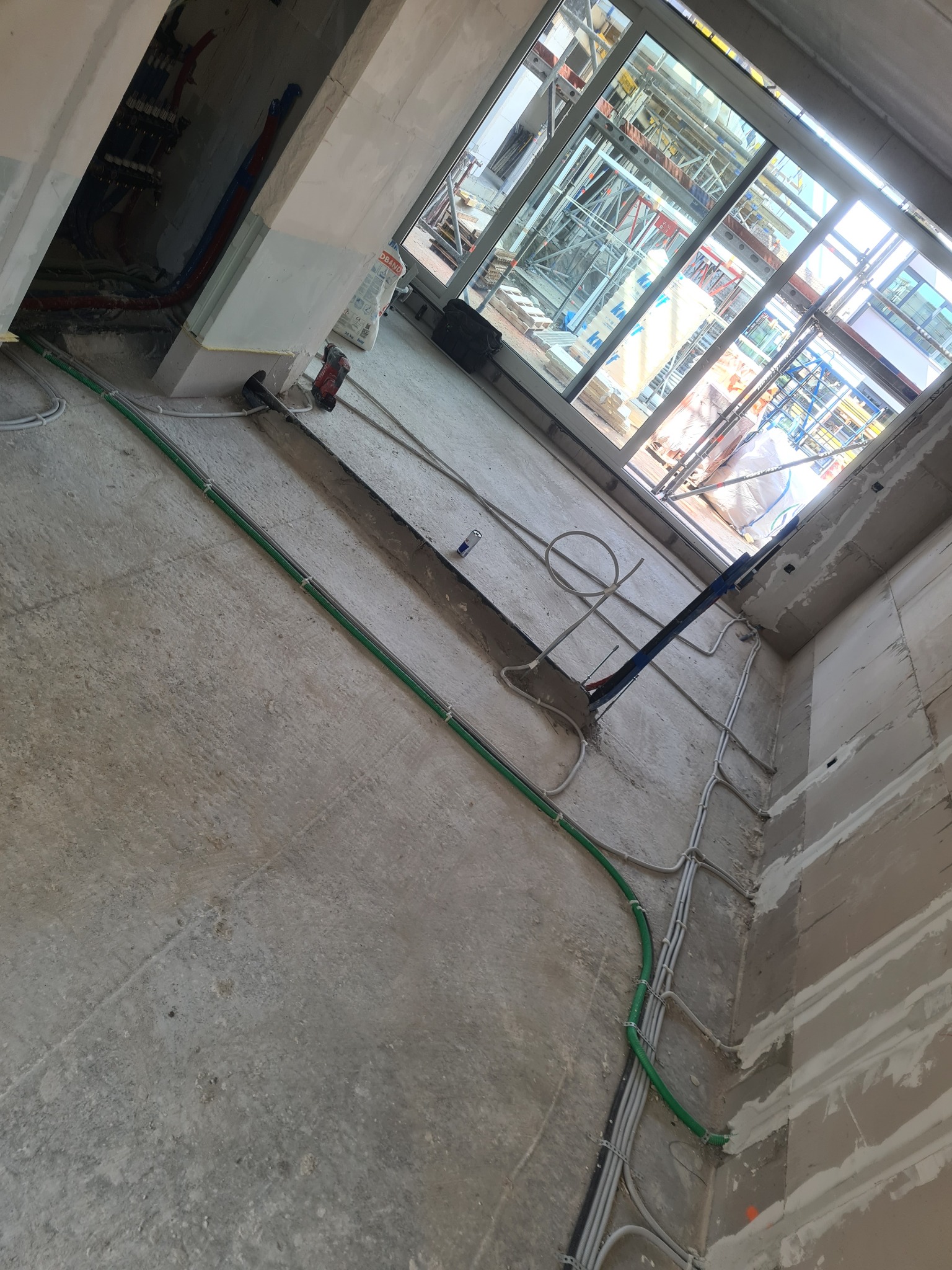 Surowy stan wnętrza w trakcie prac wykończeniowych: widoczne kable elektryczne w peszlach ułożone na podłodze i ścianach, okno z widokiem na rusztowania, ślady po gładzi na ścianach, betonowa podłoga.