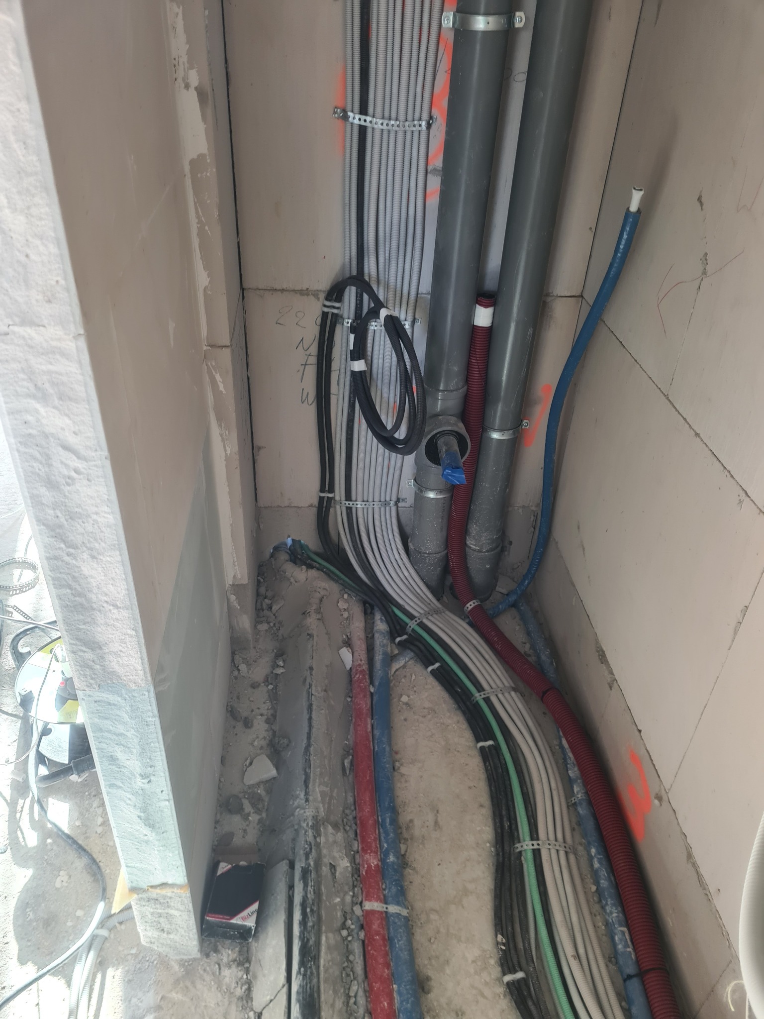 Wiązki kabli elektrycznych i rur instalacyjnych, częściowo zamocowane do ściany w trakcie montażu w surowym stanie, widoczne oznaczenia na ścianie, brak wykończenia.