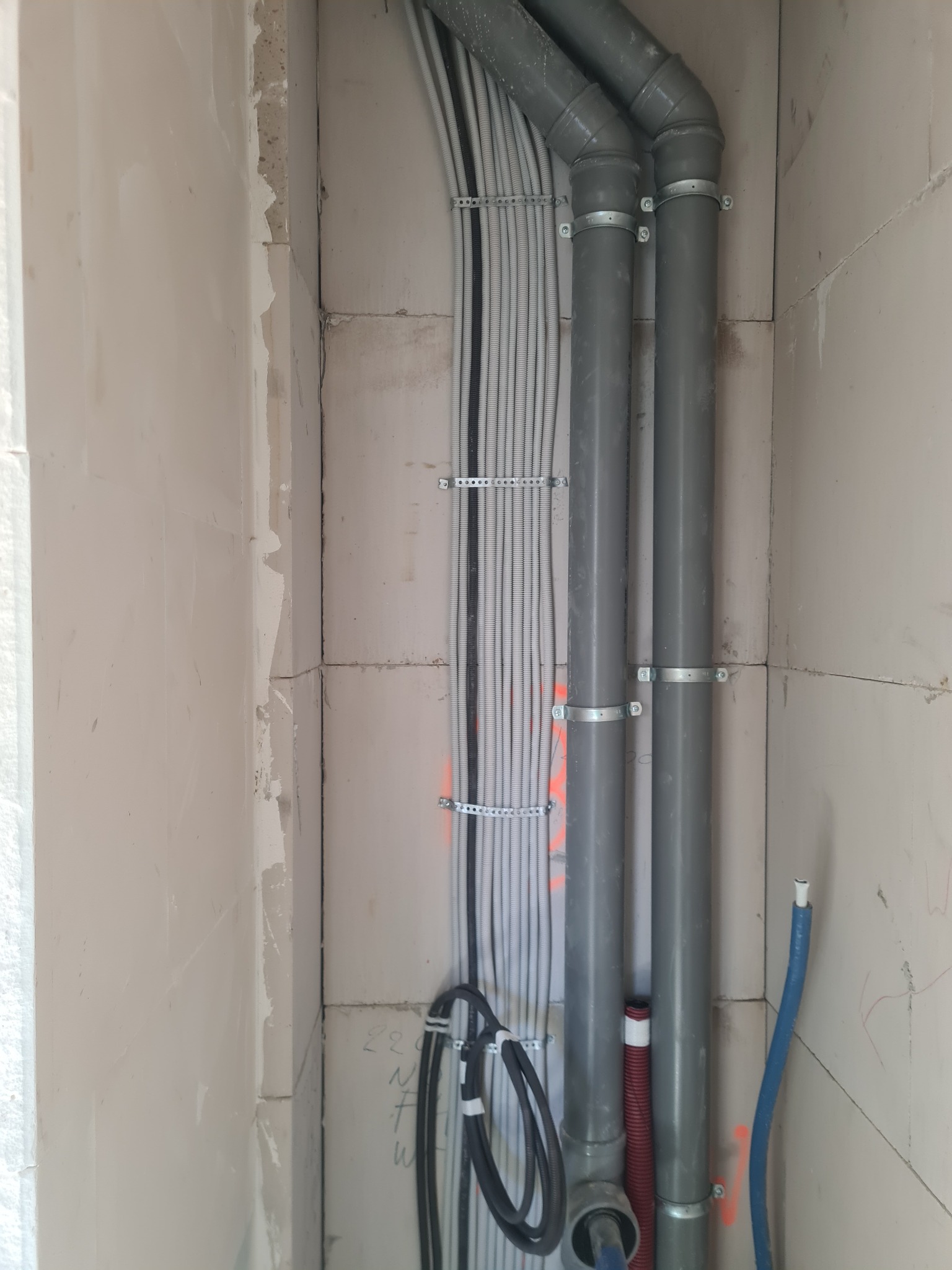 Wewnętrzne ułożenie przewodów elektrycznych i rur kanalizacyjnych w surowym stanie, przymocowanych metalowymi uchwytami do ściany z betonu komórkowego.