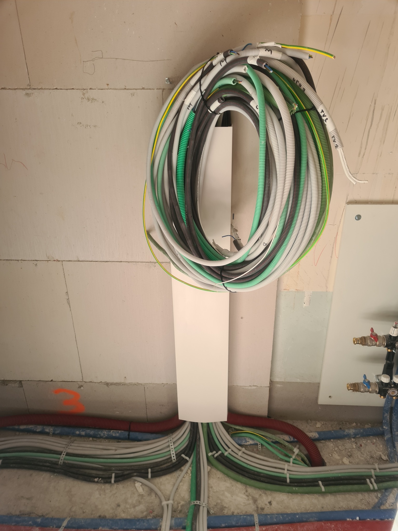 Pętle kabli elektrycznych w peszlach, zwisające z sufitu w surowym stanie, z widoczną skrzynką rozdzielczą w tle i fragmentami instalacji wodnej