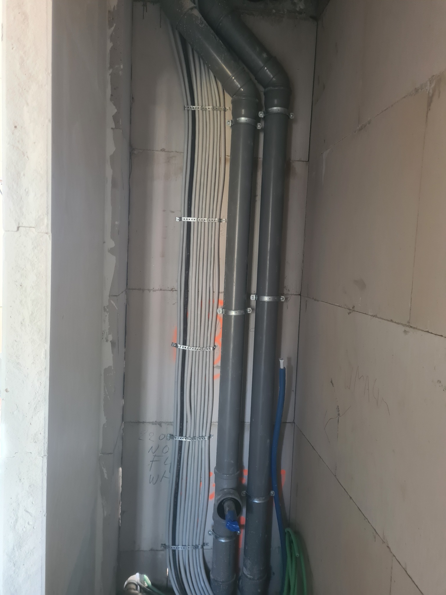 Wewnętrzny narożnik ściany z szarego betonu komórkowego, widoczne pionowe rury kanalizacyjne z szarego PCV, mocowane obejmami, oraz wiązka przewodów elektrycznych zabezpieczonych peszlami...
