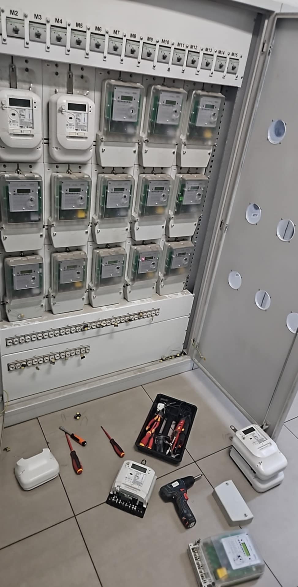 Otwarta szafa elektryczna z rzędem liczników energii, narzędziami i miernikami na podłodze. Widoczne śruby montażowe i oznaczenia M2-M15 na górze.