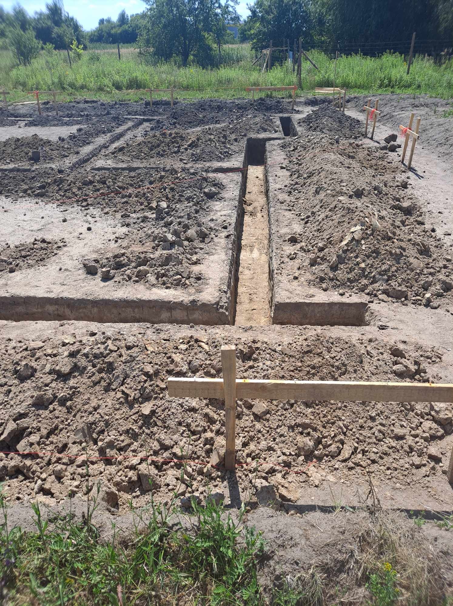 Przygotowanie terenu pod fundamenty: wykopane rowy, drewniane znaczniki z czerwoną taśmą, pryzmy ziemi, widok z góry na plac budowy.