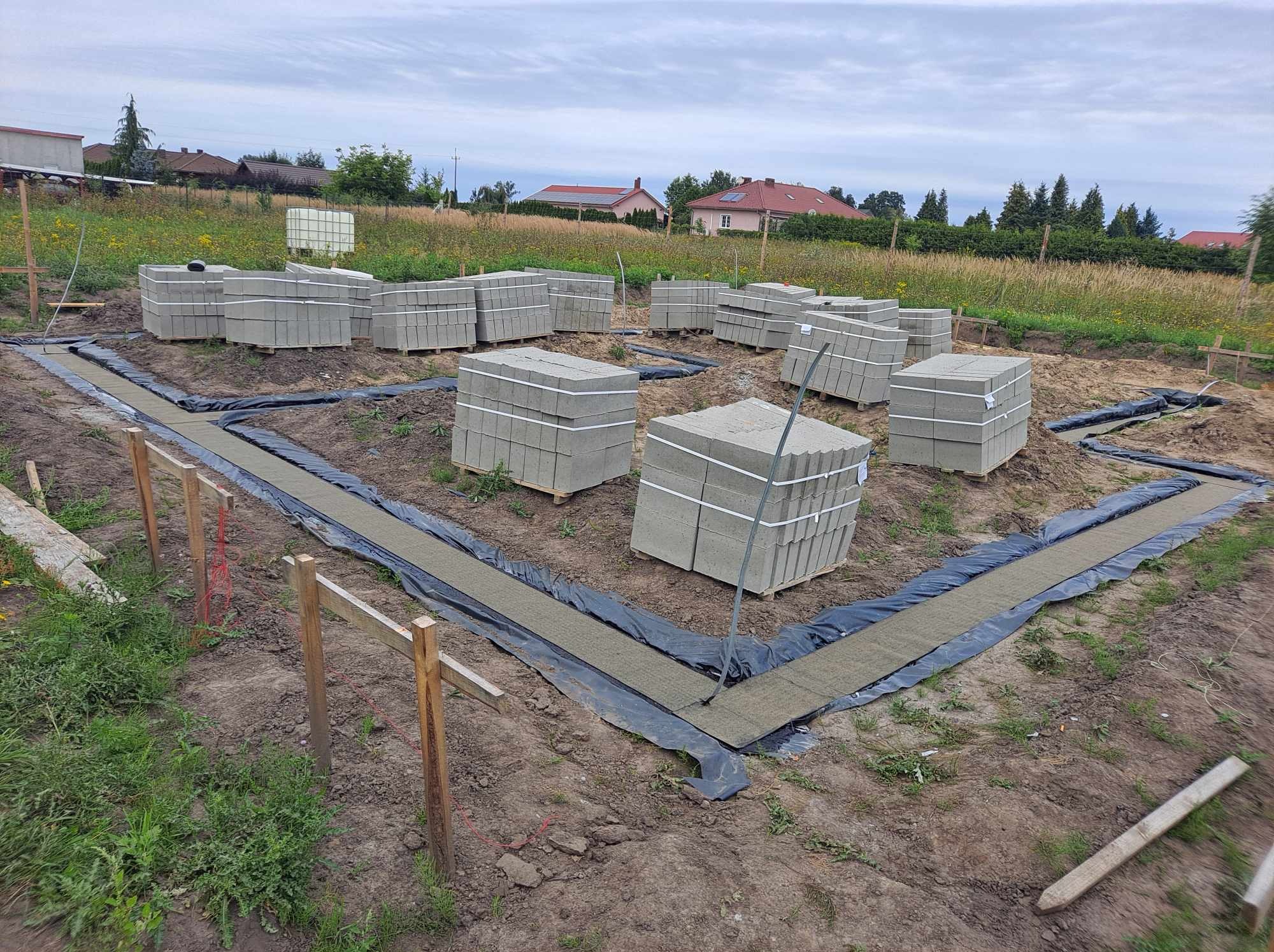 Przygotowanie terenu pod fundamenty domu: wykopane rowy wyłożone czarną folią i geowłókniną, obok ułożone palety z bloczkami betonowymi, widoczne drewniane szalunki i otaczająca roślinność.