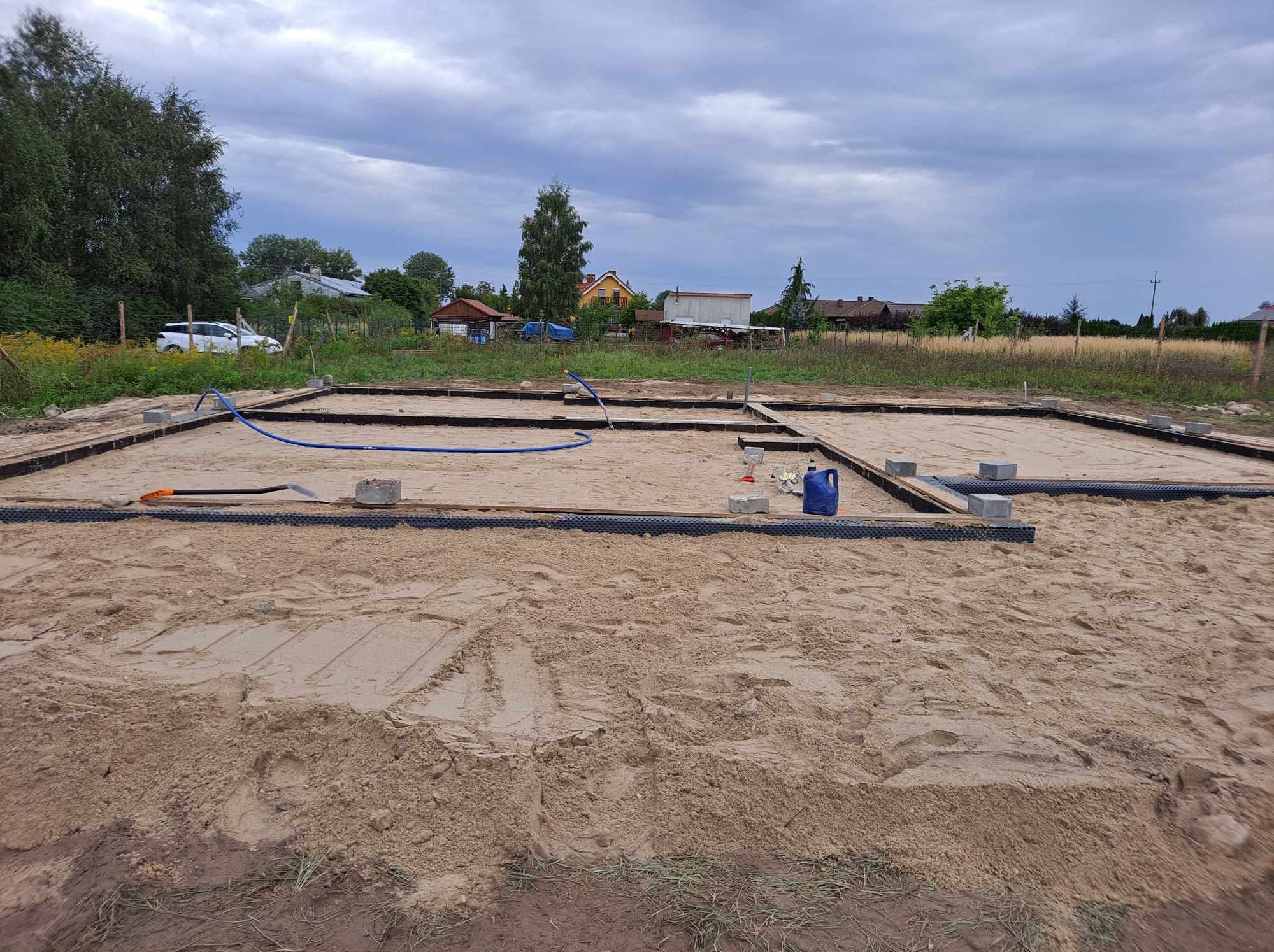 Przygotowanie terenu pod fundamenty domu jednorodzinnego: wytyczone obrysy, piasek, obrzeża z tworzywa, betonowe bloczki, niebieski wąż i narzędzia w tle wiejskich zabudowań pod pochmurnym niebem.