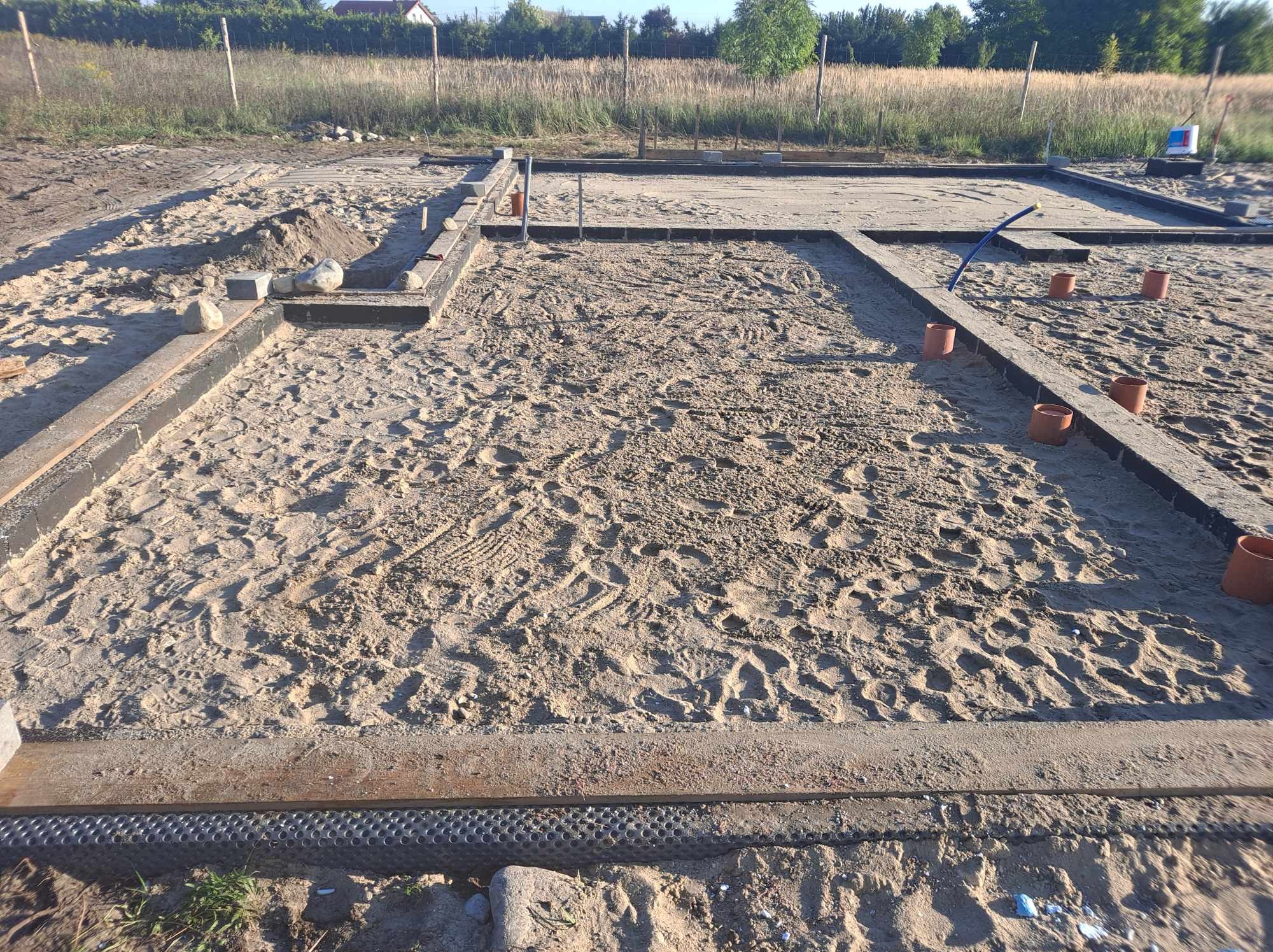 Przygotowany teren pod budowę domu z widocznymi fundamentami, zasypanymi piaskiem i zaznaczonymi rurami kanalizacyjnymi.