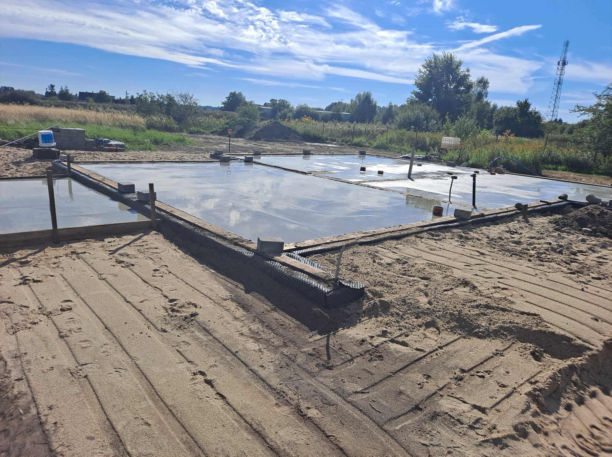 Wylana płyta fundamentowa z widocznym zbrojeniem, deskowaniem i instalacjami, otoczona piaskiem na tle krajobrazu z drzewami i błękitnym niebem.
