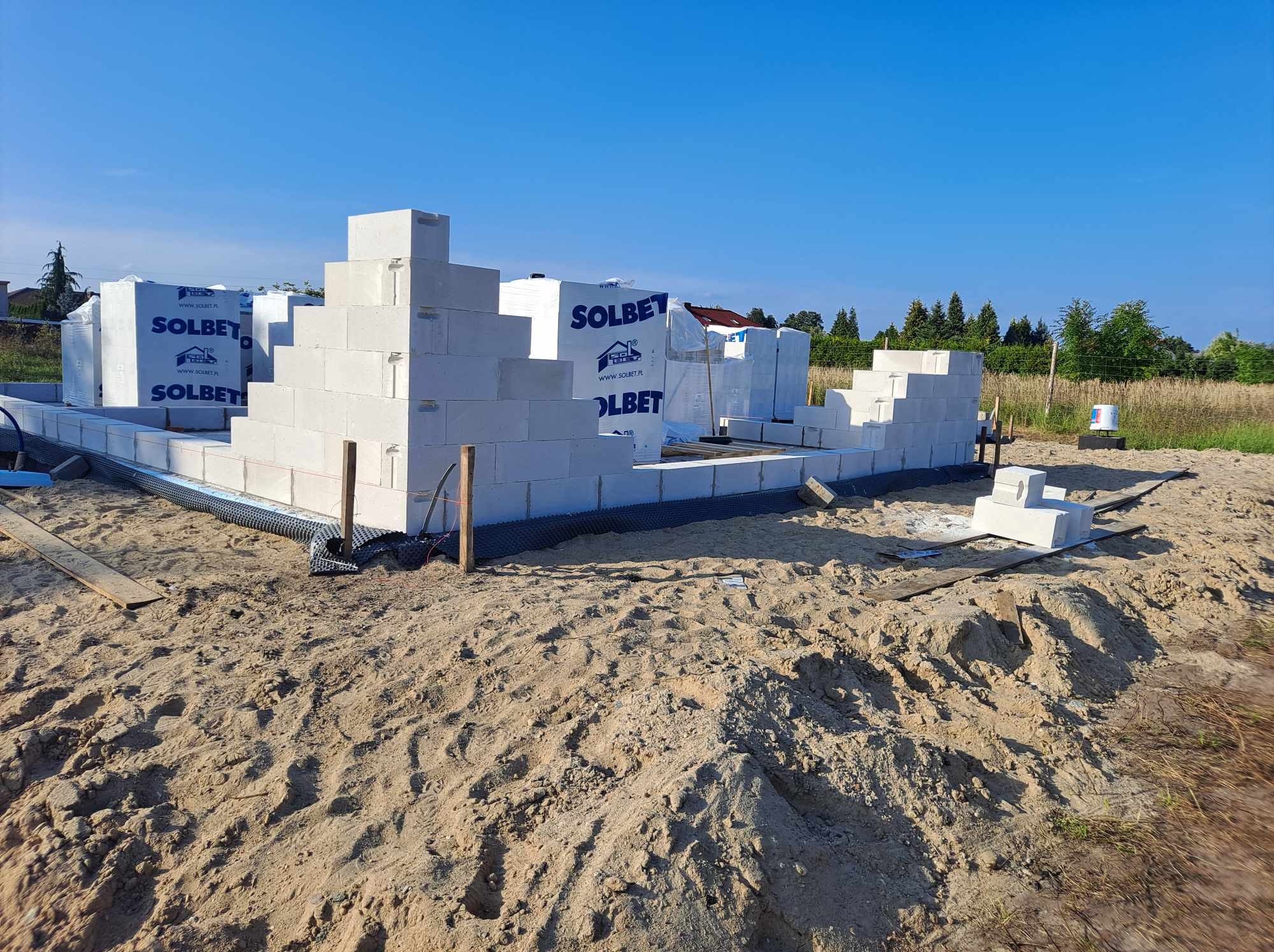 Wznoszenie muru z białych bloczków Solbet na fundamencie z widoczną izolacją przeciwwilgociową, w tle zapakowane bloczki, teren piaszczysty.