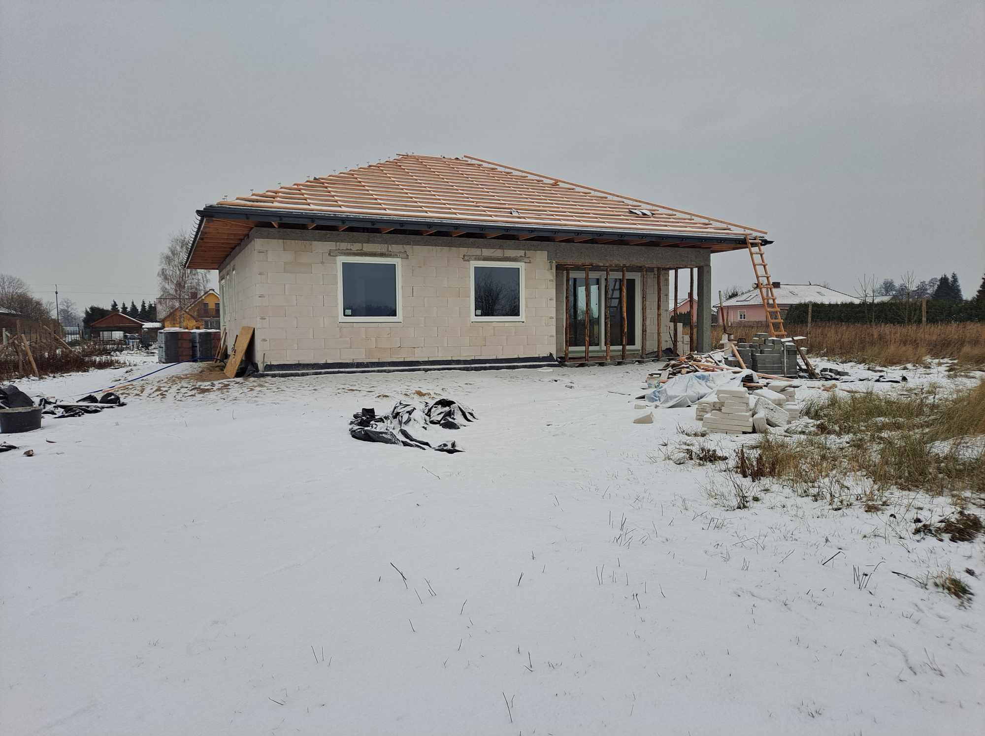 Surowy stan domu jednorodzinnego w zimowej scenerii, widoczne białe bloczki, drewniana konstrukcja dachu z dachówką, okna bez parapetów, drabina oparta o ścianę, śnieg na ziemi i pochmurne niebo.