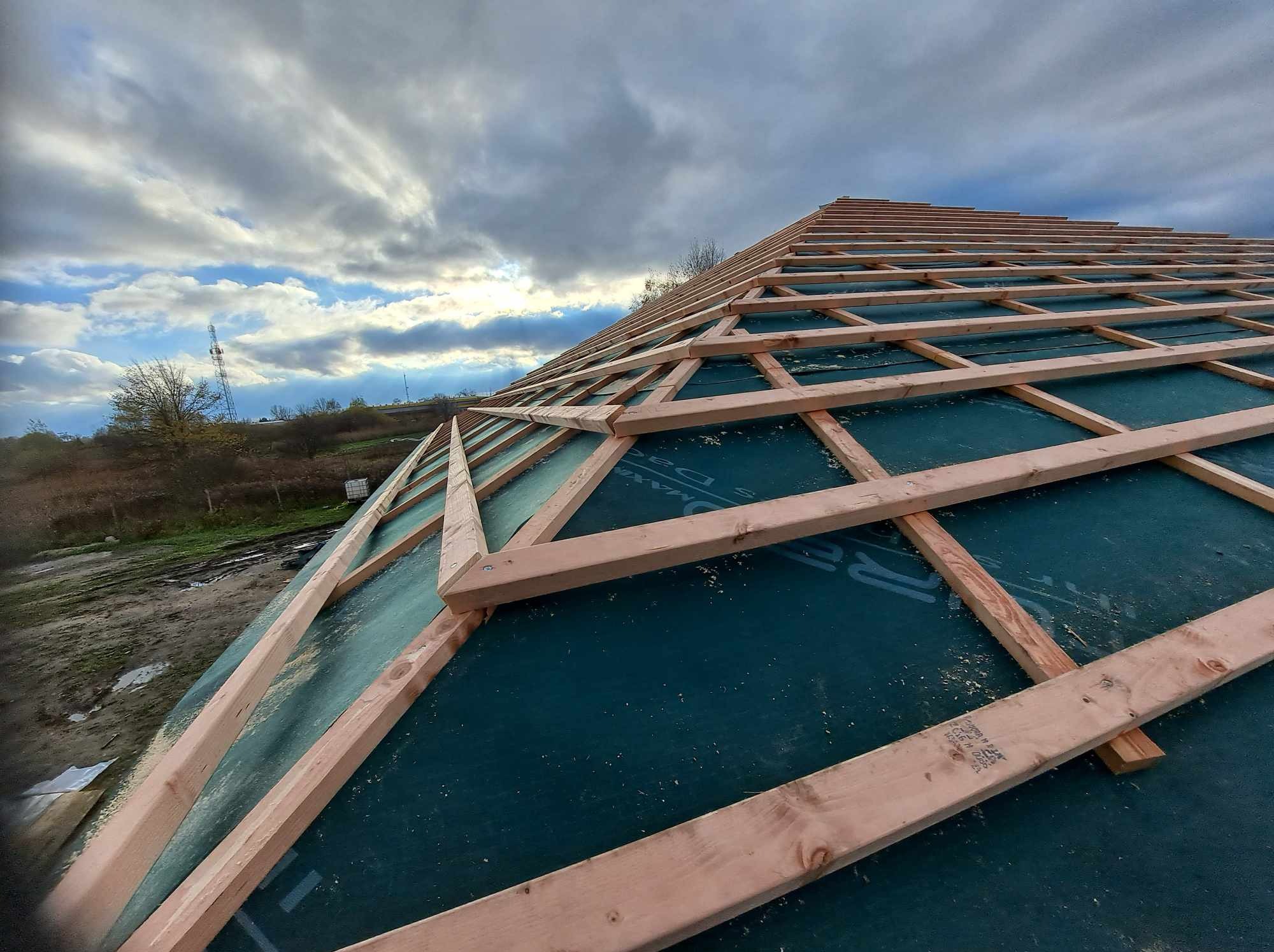 Konstrukcja dachu z drewnianych łat i papy w trakcie budowy, widok z dołu na tle pochmurnego nieba i krajobrazu z masztem telekomunikacyjnym.
