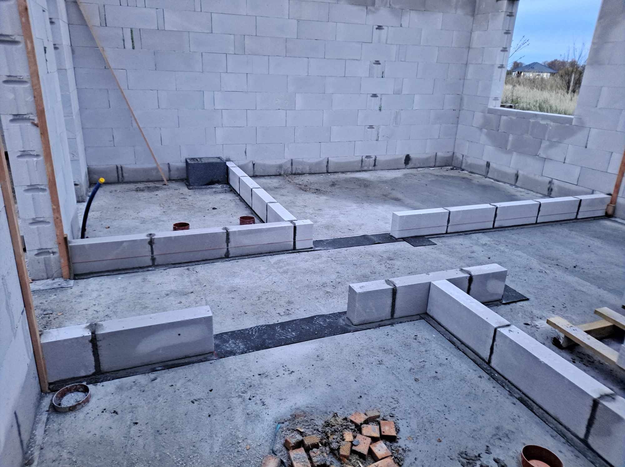 Wnętrze w surowym stanie z wymurowanymi ścianami z betonu komórkowego, przygotowane do dalszych prac wykończeniowych, widoczne fragmenty instalacji i resztki materiałów budowlanych na podłodze.